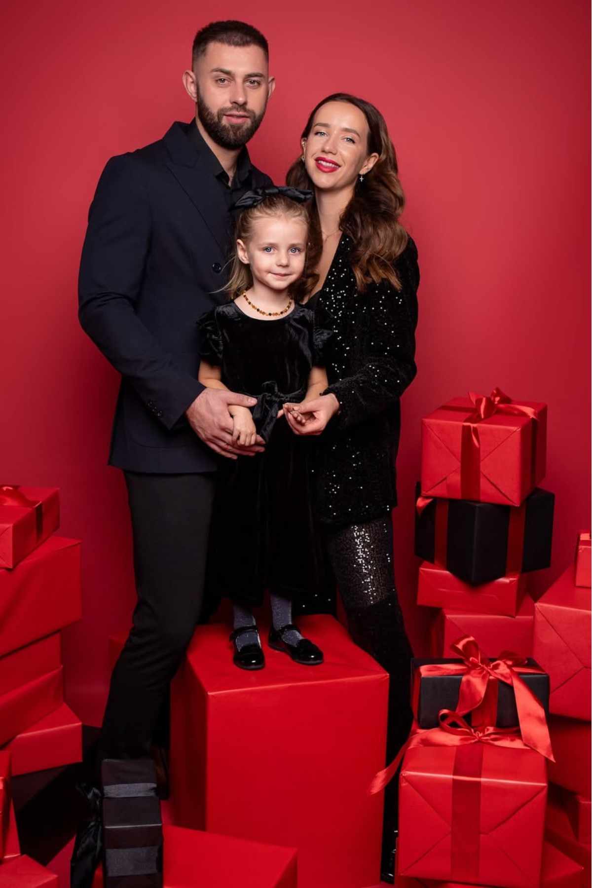 A family of three, dressed in black, poses together among stacked red and black gift boxes against a red background.