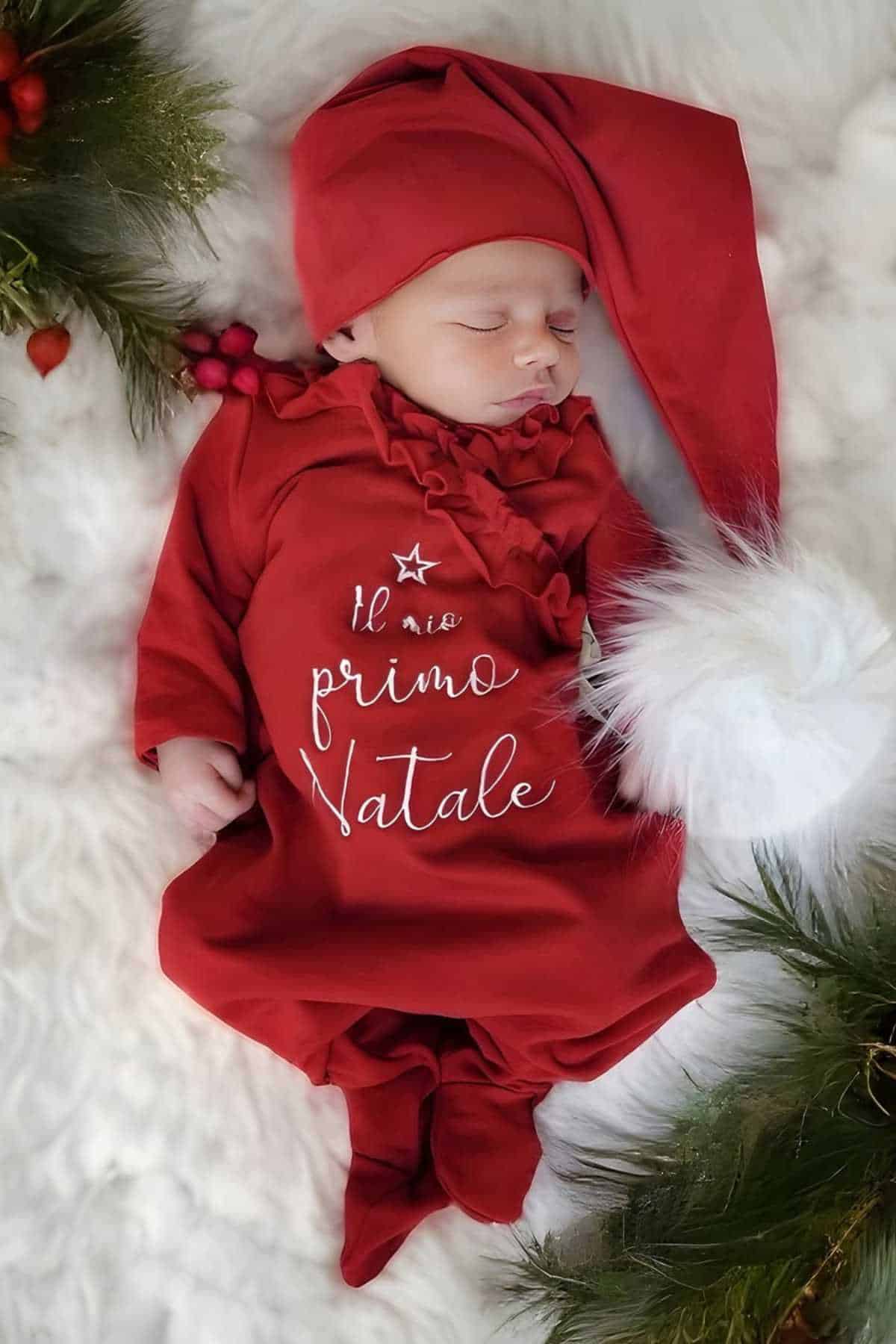 A baby in a red outfit and hat sleeps on a white blanket surrounded by greenery. The outfit reads "Il mio primo Natale," which means "My first Christmas" in Italian.