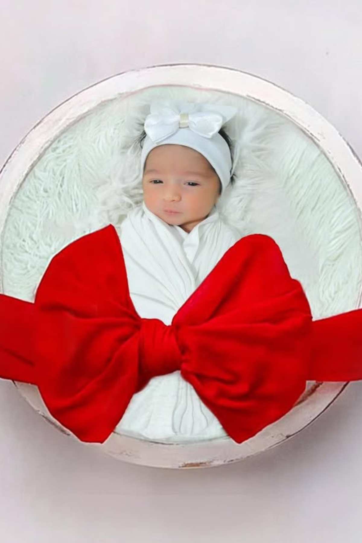 A newborn baby wrapped in a white blanket with a white headband, lying on a furry white cushion, and decorated with a large red bow.