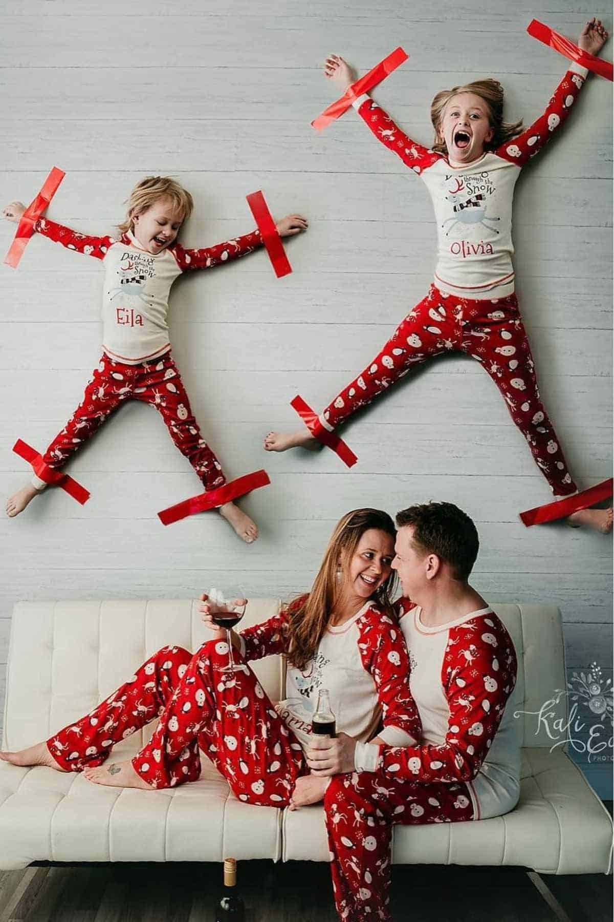 Two children in matching pajamas are taped to a wall with red tape, while two adults in matching pajamas sit on a couch below, smiling and holding drinks.