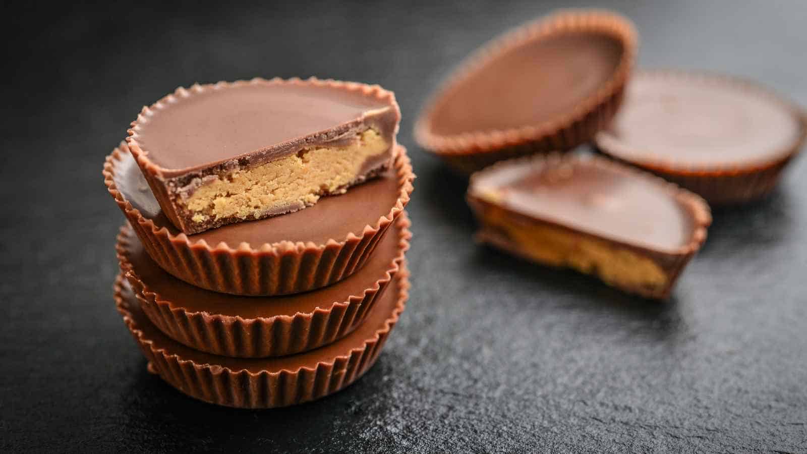 A stack of chocolate peanut butter cups is shown, with one cup cut in half to reveal the peanut butter filling. Two more halves are in the background on a dark surface.