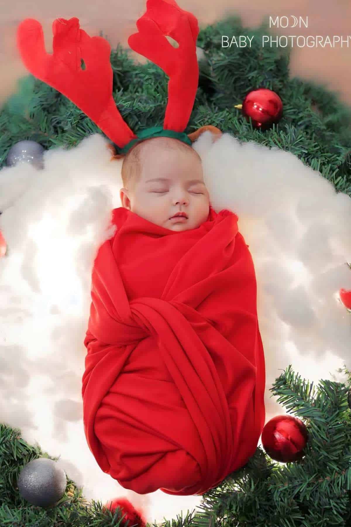 A baby wrapped in a red blanket, wearing antler headbands, sleeps on white padding surrounded by Christmas ornaments and greenery.