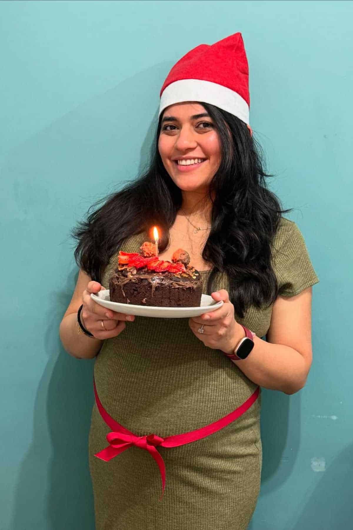 A woman wearing a Santa hat holds a chocolate cake with a lit candle, smiling in front of a blue wall.