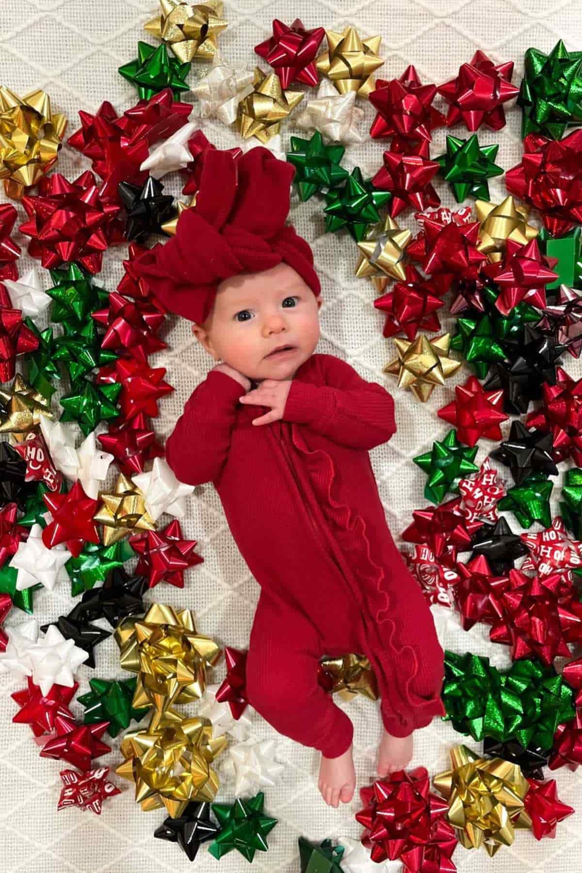 A baby in a red outfit and matching headband lies on a white surface surrounded by red, green, gold, and white gift bows.