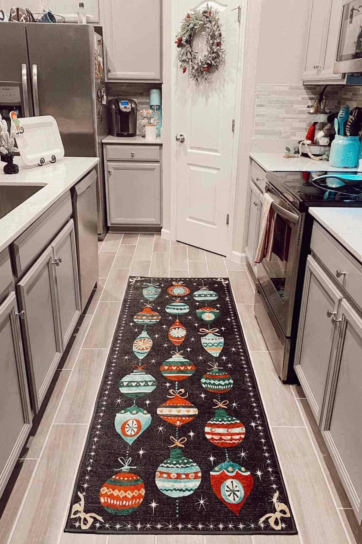 A kitchen with gray cabinets and appliances decorated for the holidays, featuring a rug with colorful ornament designs on the floor.