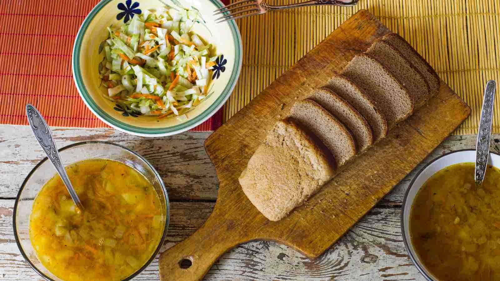 A wooden table with two bowls of soup, a bowl of shredded cabbage salad, and sliced brown bread on a wooden cutting board.
