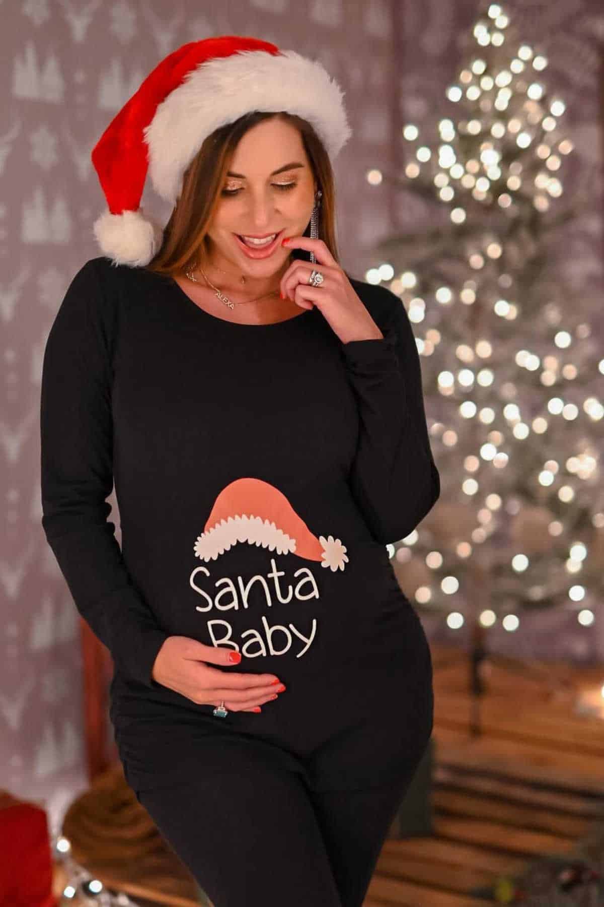 A pregnant woman wearing a Santa hat and a black "Santa Baby" outfit stands indoors near a blurred, lit Christmas tree.