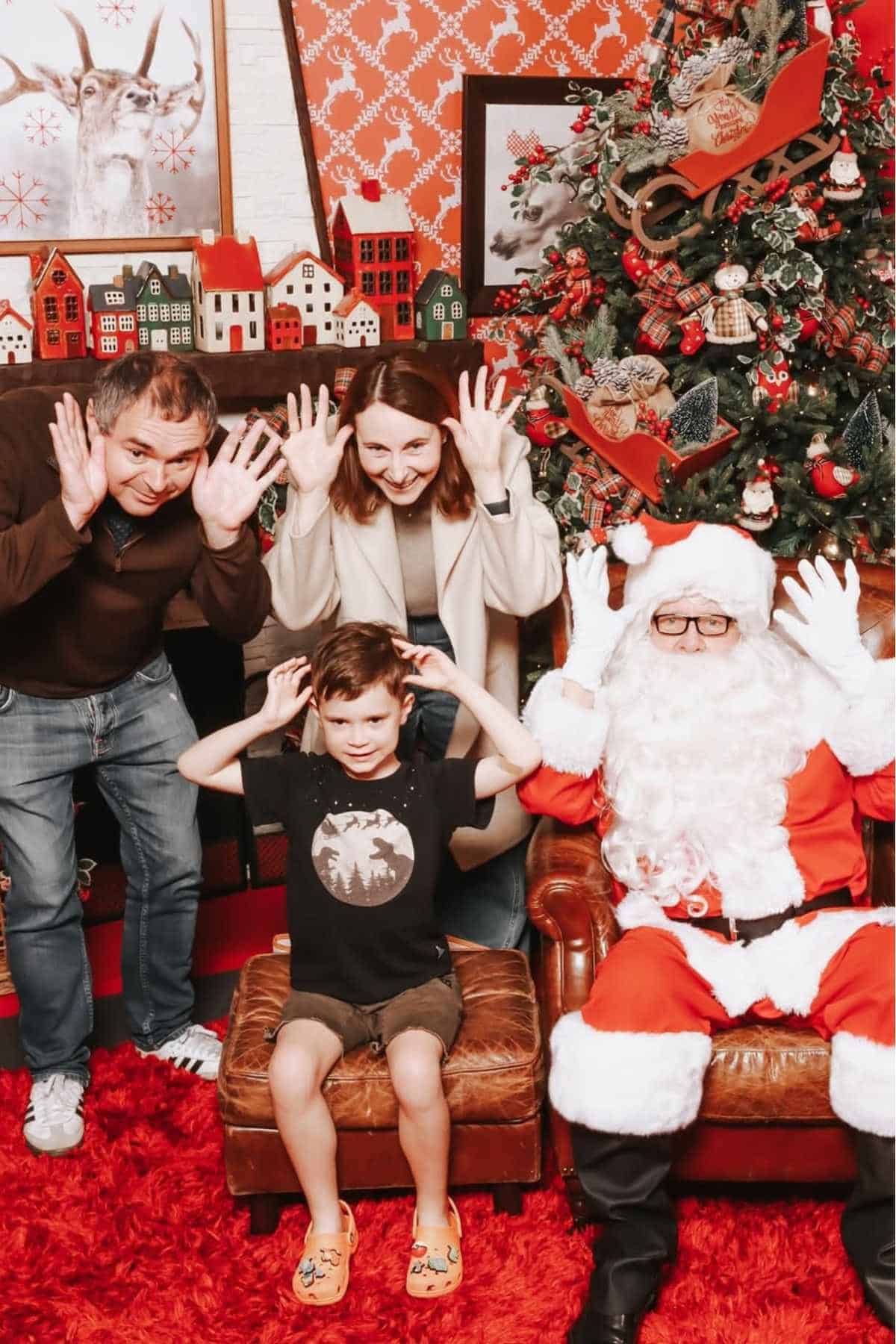 Four people, including a child and a person dressed as Santa Claus, pose playfully with raised hands in front of a decorated Christmas tree.
