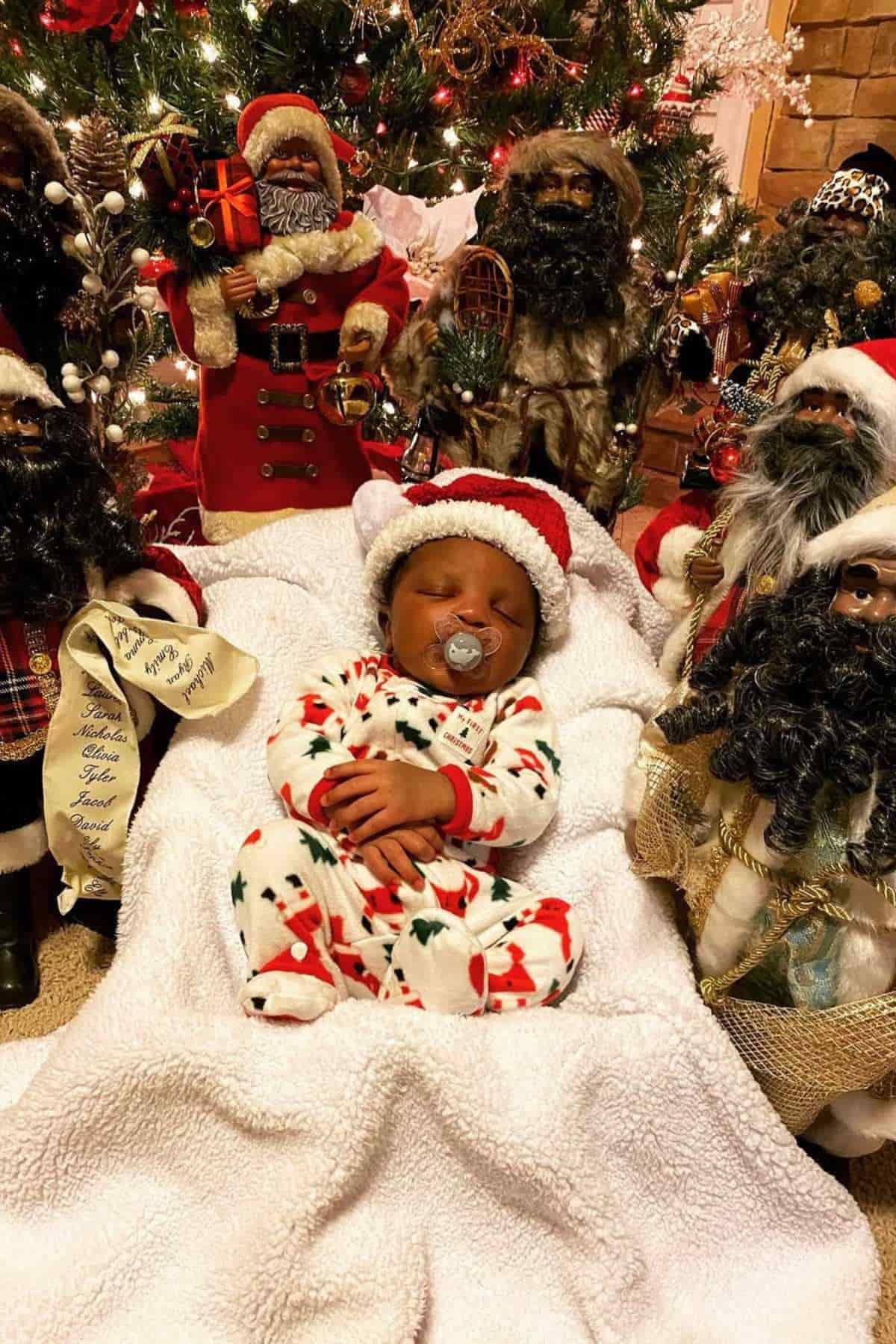A baby in Christmas pajamas and a Santa hat sleeps on a white blanket, surrounded by several Black Santa Claus figurines in front of a decorated Christmas tree.