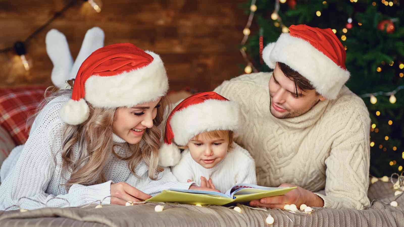A family wearing Santa hats lies on the floor next to a Christmas tree, reading a book together and surrounded by festive lights.