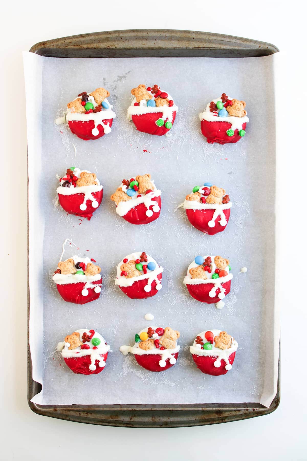 A baking tray with twelve red cupcakes topped with white frosting, cookies, and colorful candies, on parchment paper.