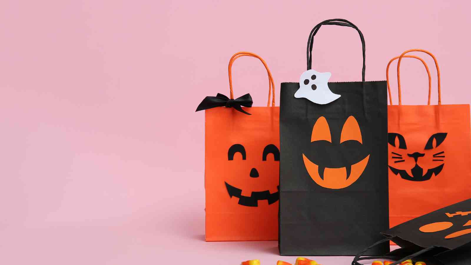 Three Halloween-themed gift bags with jack-o'-lantern and cat faces on a pink background; one black bag has a ghost decoration.