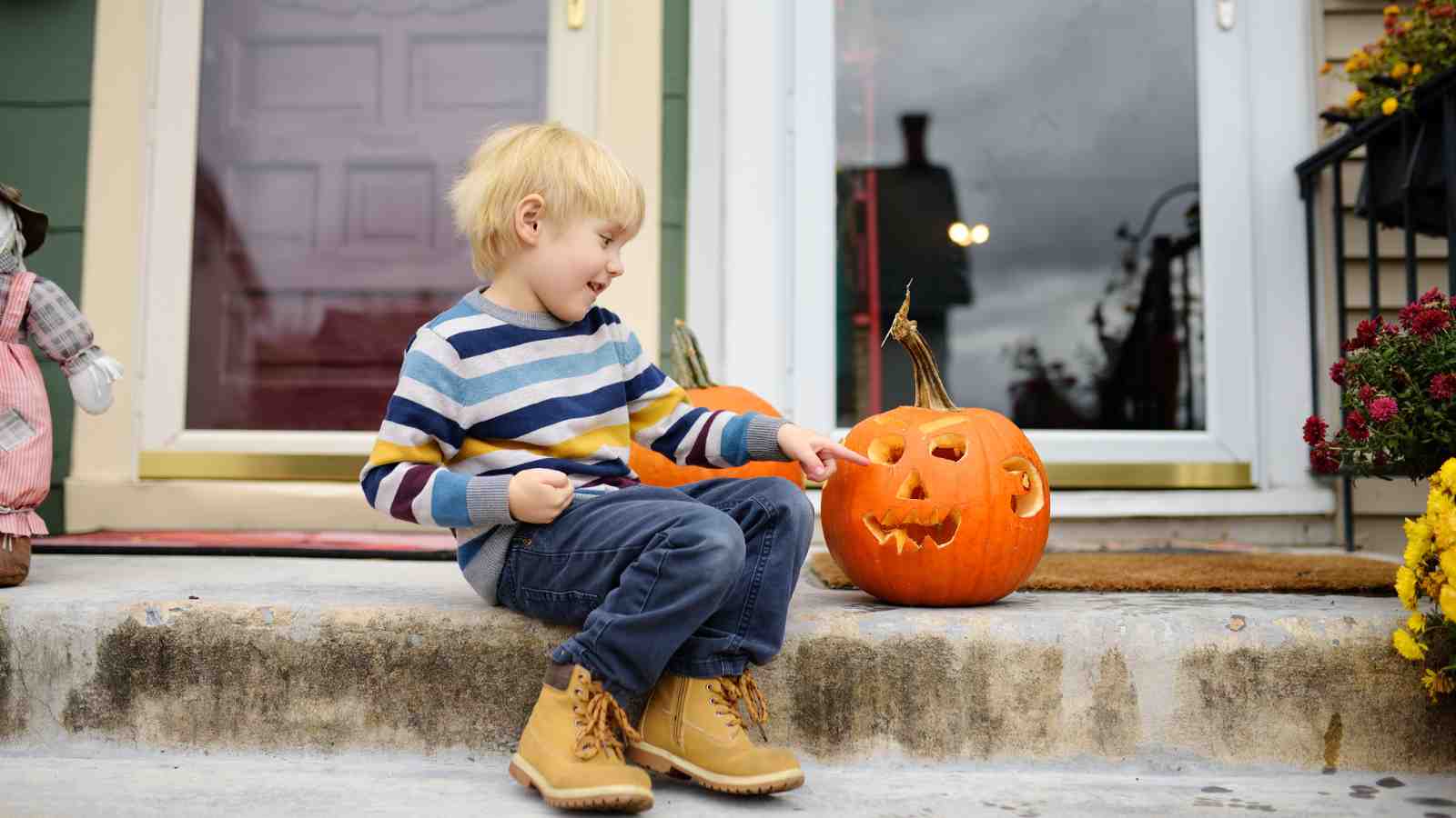 A young child in a striped sweater sits on a porch step, smiling and pointing at a carved jack-o’-lantern pumpkin beside them.