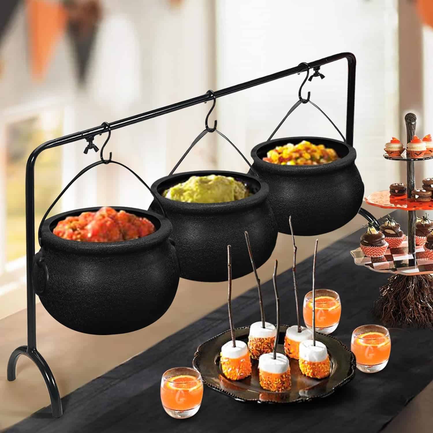 Three black cauldrons filled with food hang on a metal stand over a table set with drinks, marshmallow treats, and cupcakes.