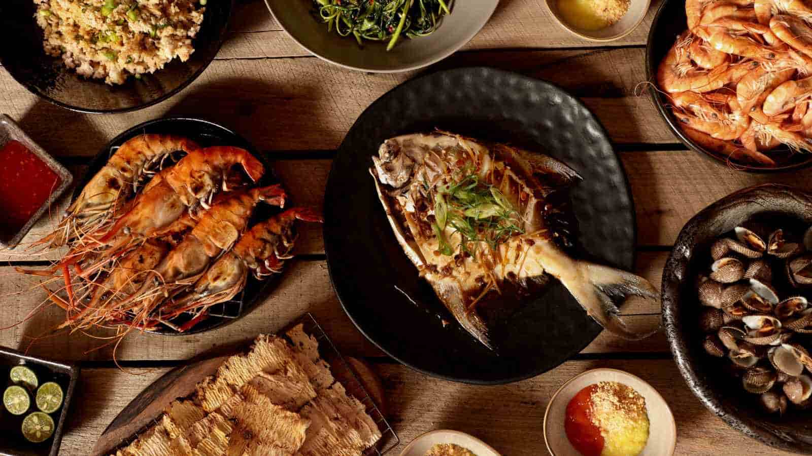 A variety of seafood dishes including grilled fish, prawns, clams, and sides are arranged on a wooden table.