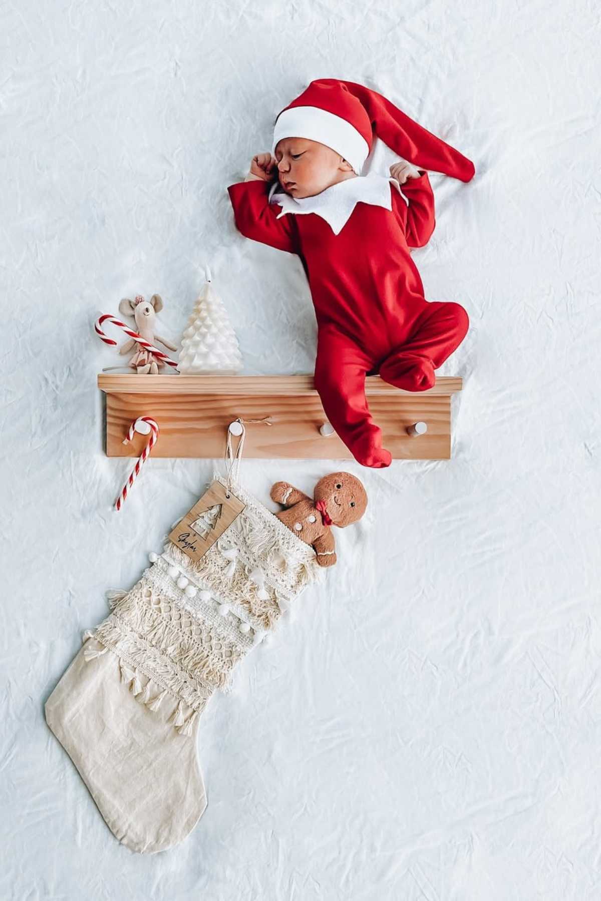 A baby dressed in a red elf costume sleeps on a wooden shelf, with Christmas decorations and a large stocking below on a white background.
