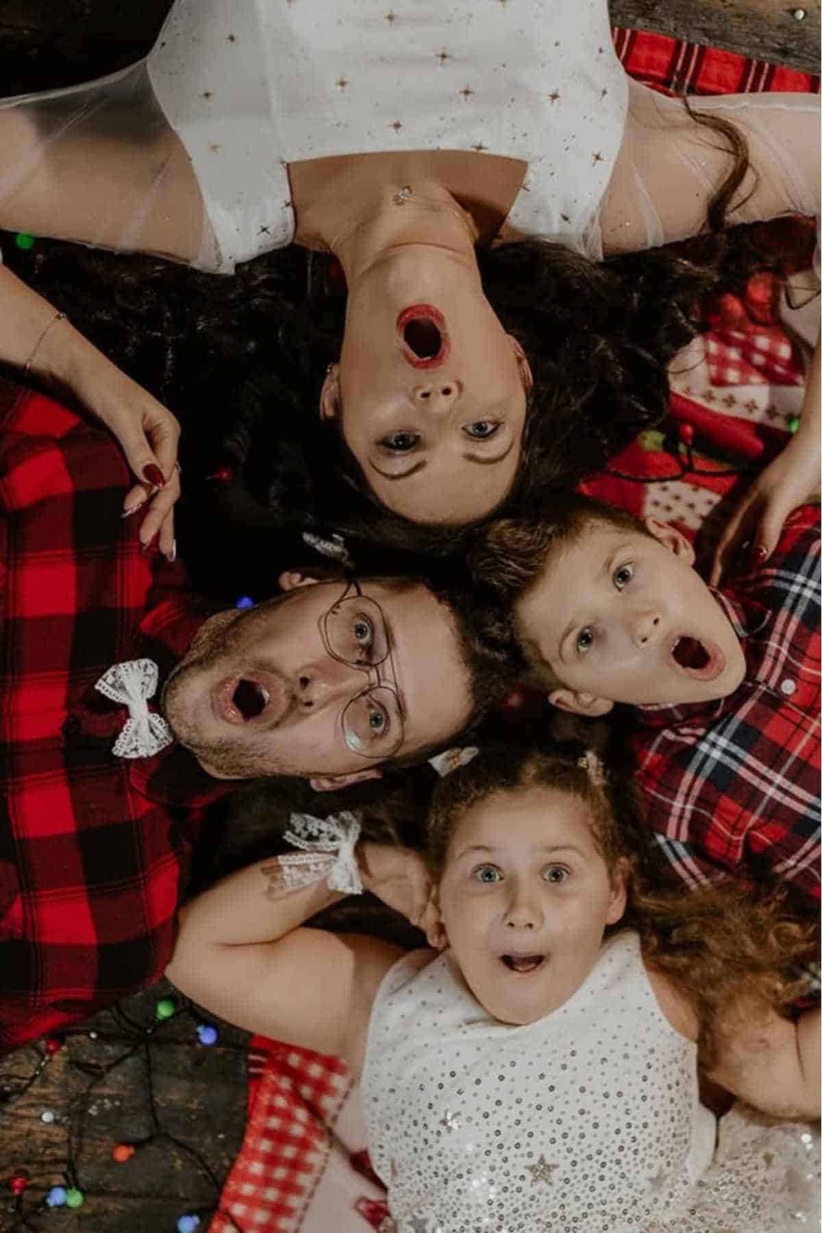 Four people, two adults and two children, lie on the floor in a circle with surprised expressions, surrounded by festive decorations and lights.