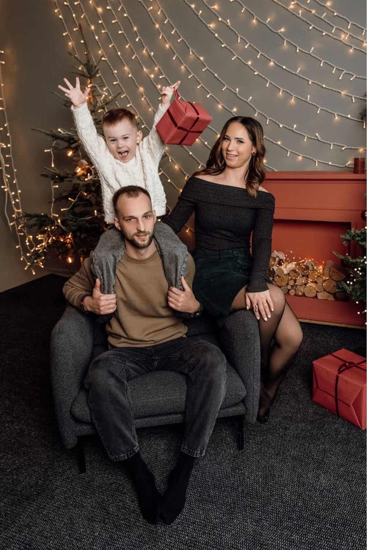A man sits in a chair with a child on his shoulders; a woman sits on the chair’s arm. They are surrounded by Christmas decorations and lights.