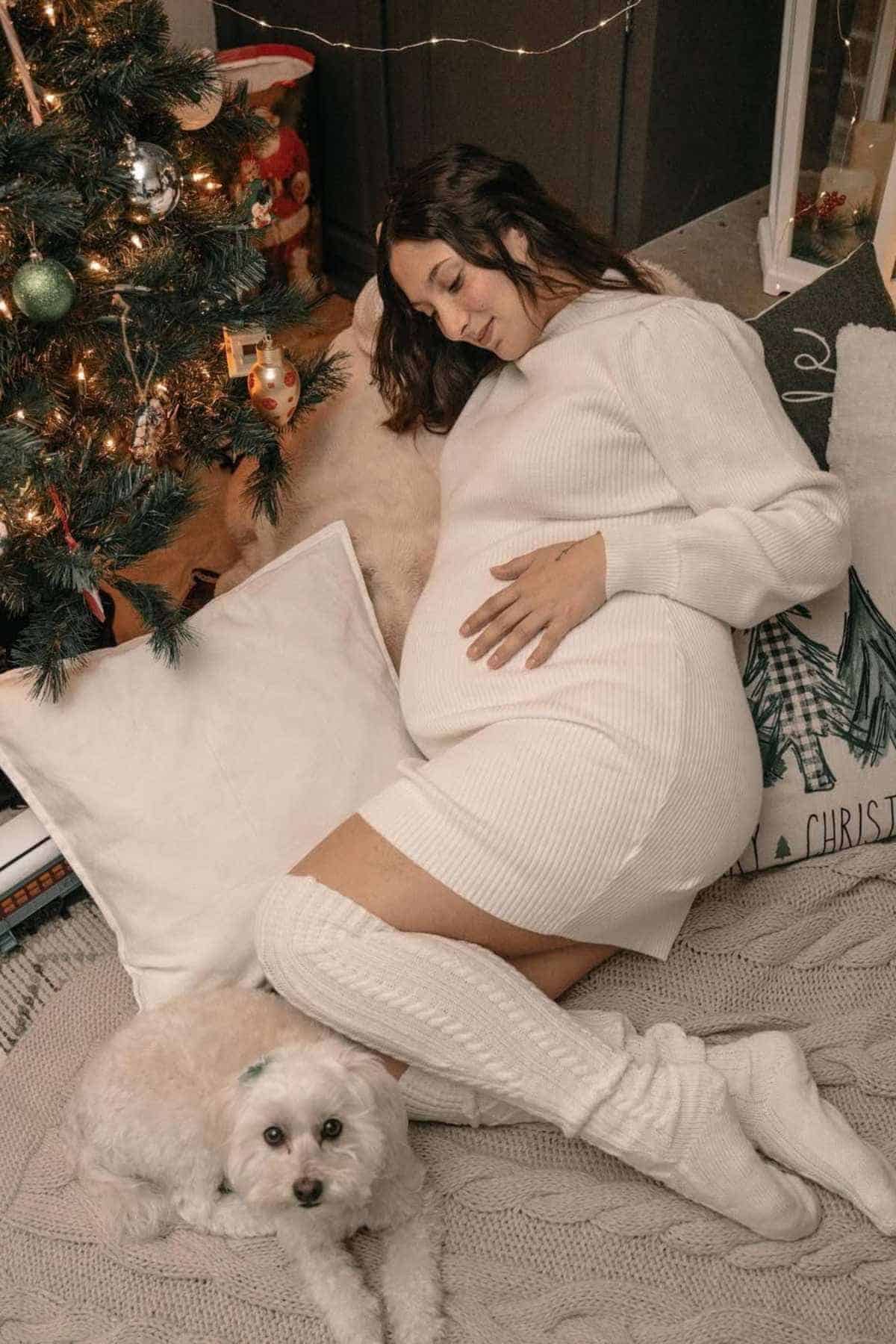 Pregnant woman in a white dress lies on a blanket near a Christmas tree, resting her hand on her belly, with a small white dog lying beside her.