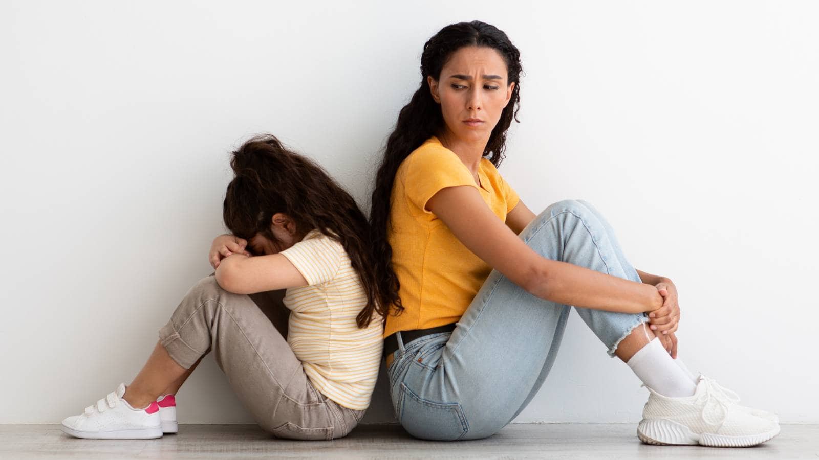 An adult woman and a young girl sit back to back on the floor against a white wall, both appearing upset and not looking at each other.
