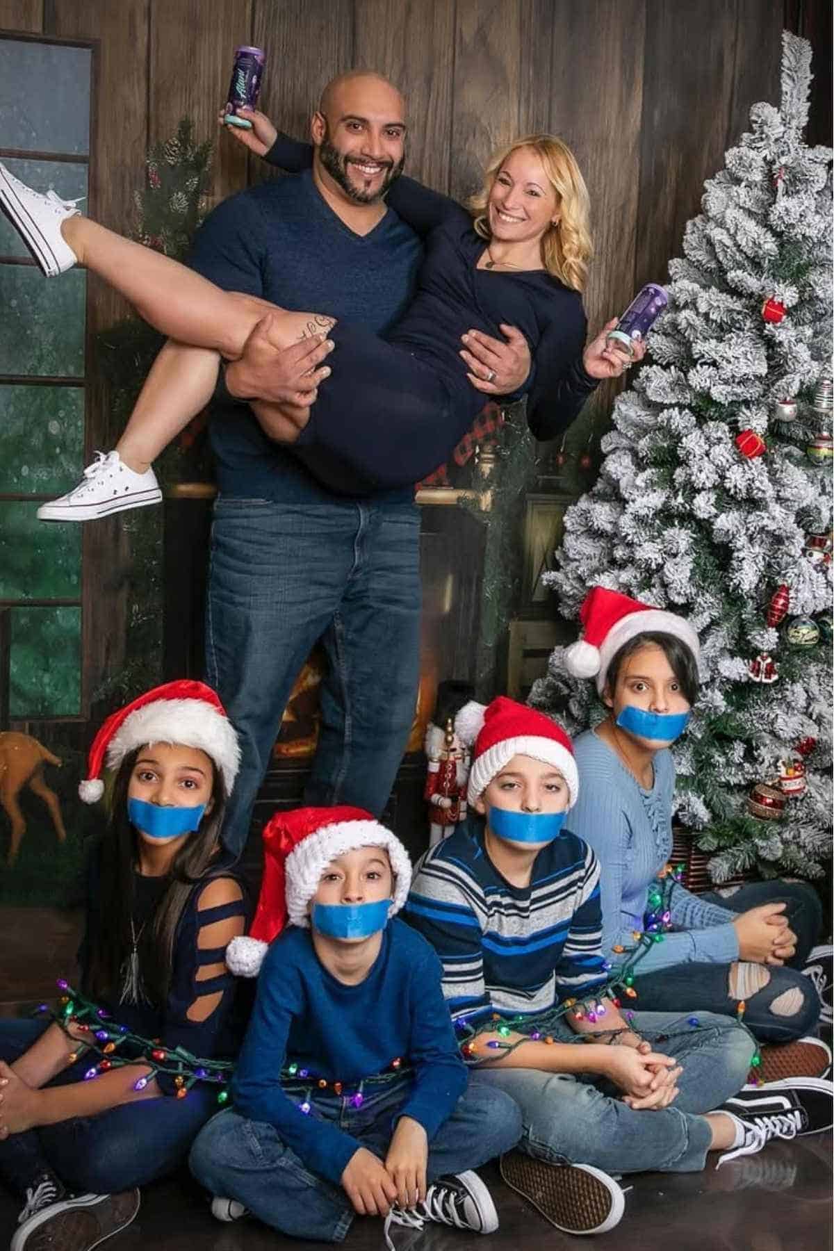 A man holds a woman while four children wearing Santa hats and blue tape over their mouths sit in front of a decorated Christmas tree.
