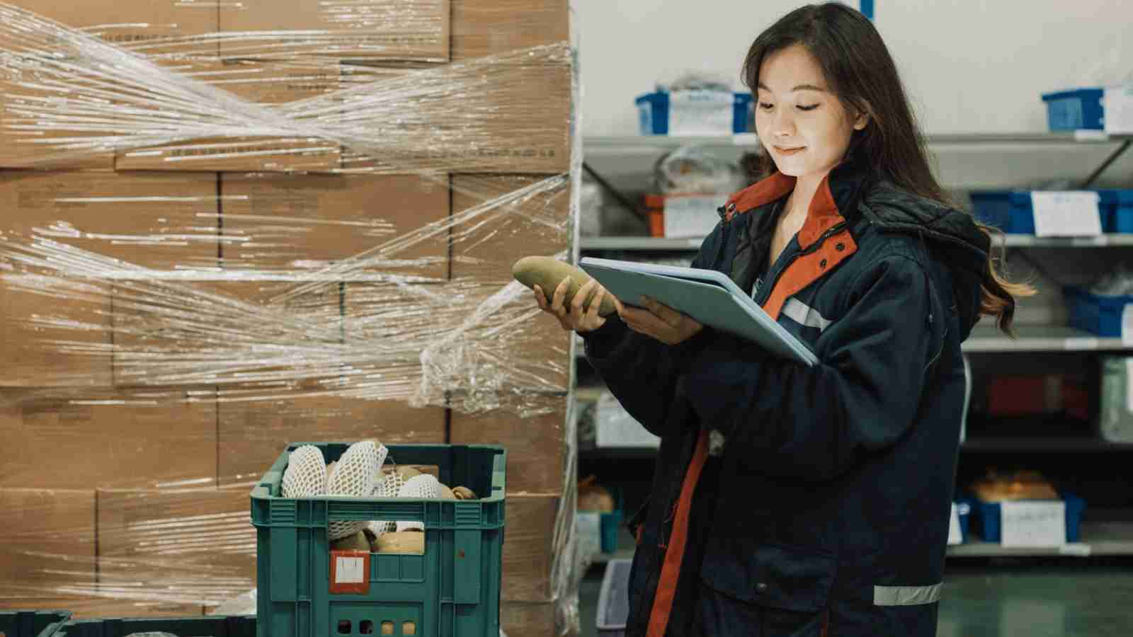 A woman in a warehouse inspects produce with a tablet, standing beside crates of packaged vegetables—an approach seen in countries that handle food waste better than the US.