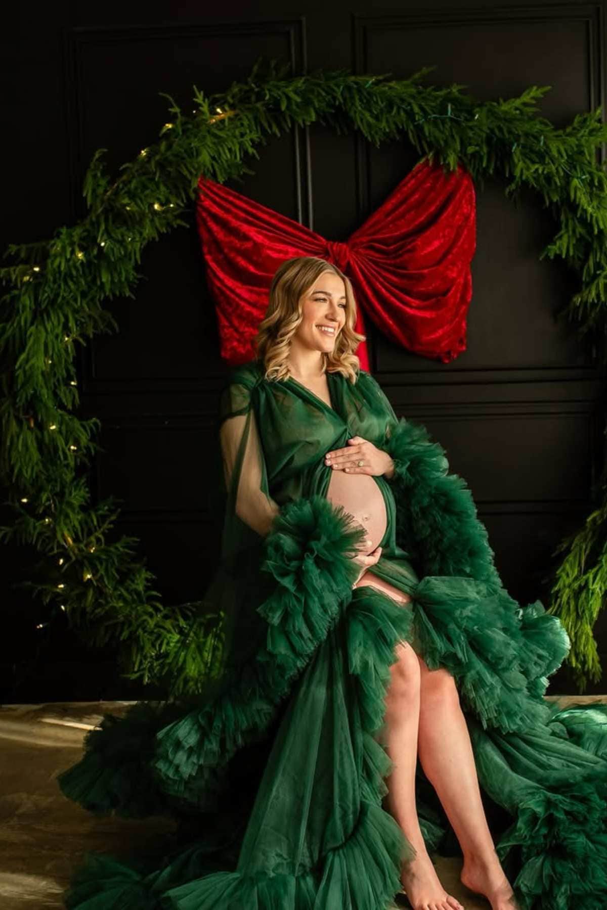 A pregnant woman in a green gown sits barefoot, smiling and holding her belly, with a holiday wreath and red bow behind her.