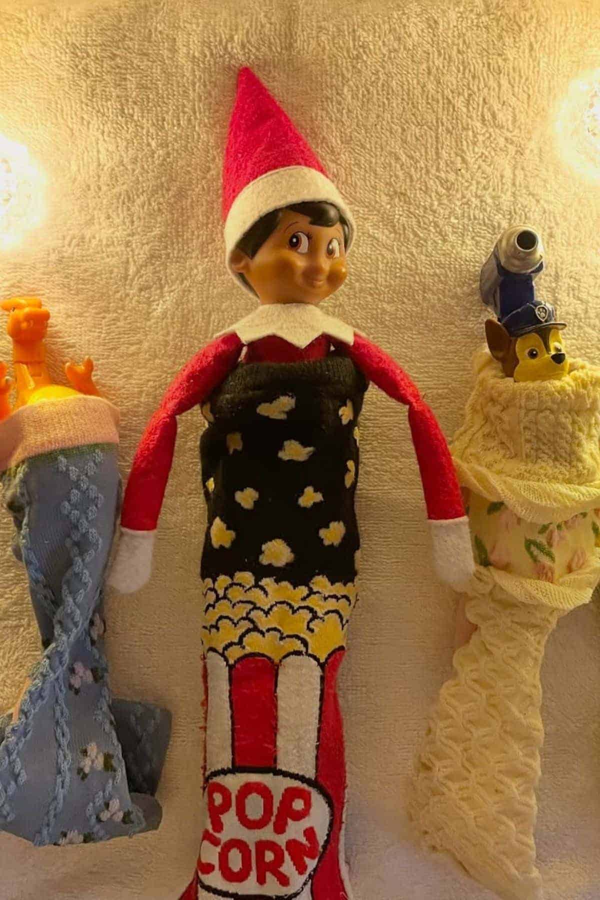 Three toy figures wrapped in patterned cloth stand upright against a light background; the center figure is dressed as an elf with a popcorn design.