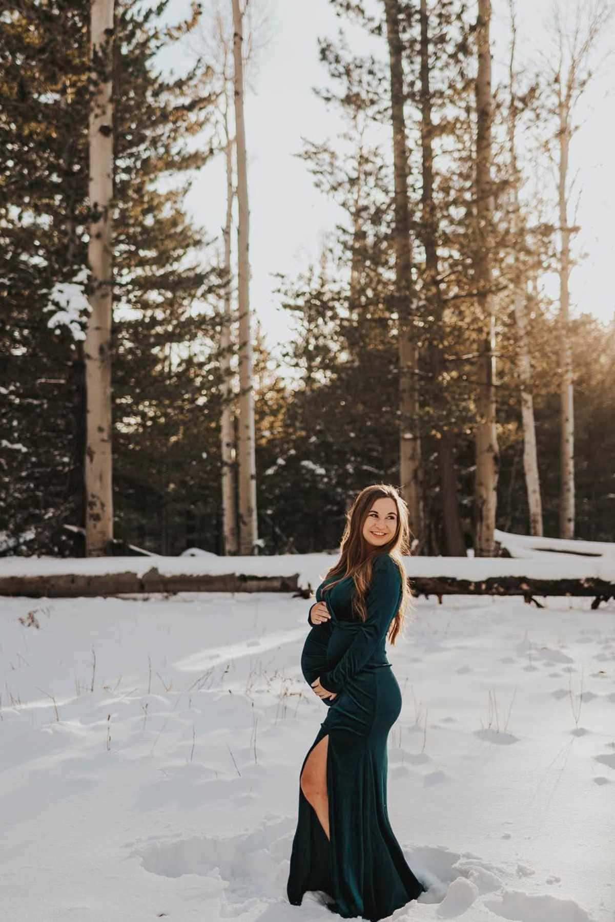 A woman in a dark green dress stands in the snow in a forest, holding her baby bump and smiling at the camera. Sunlight filters through the trees in the background.