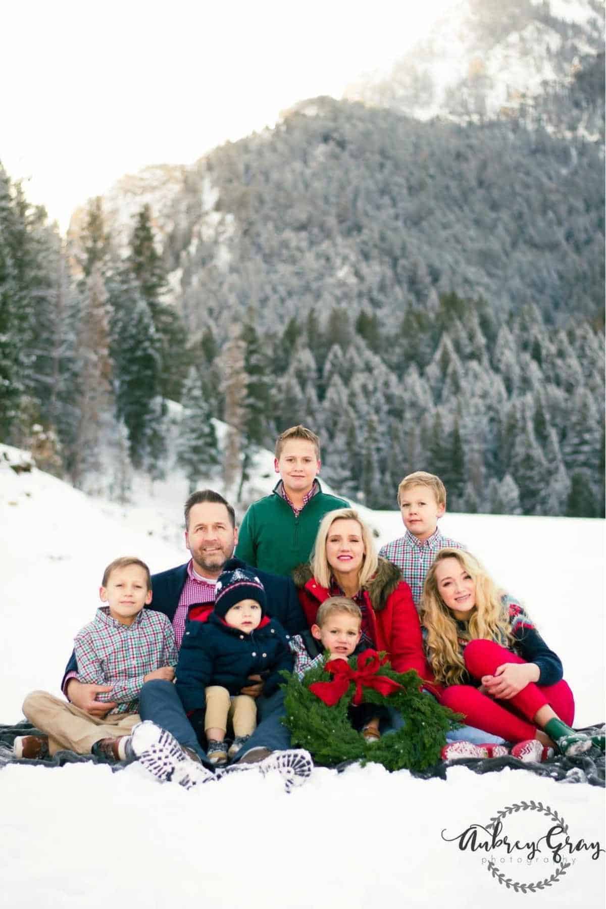 A family of eight poses outdoors in a snowy landscape with pine trees and mountains in the background.
