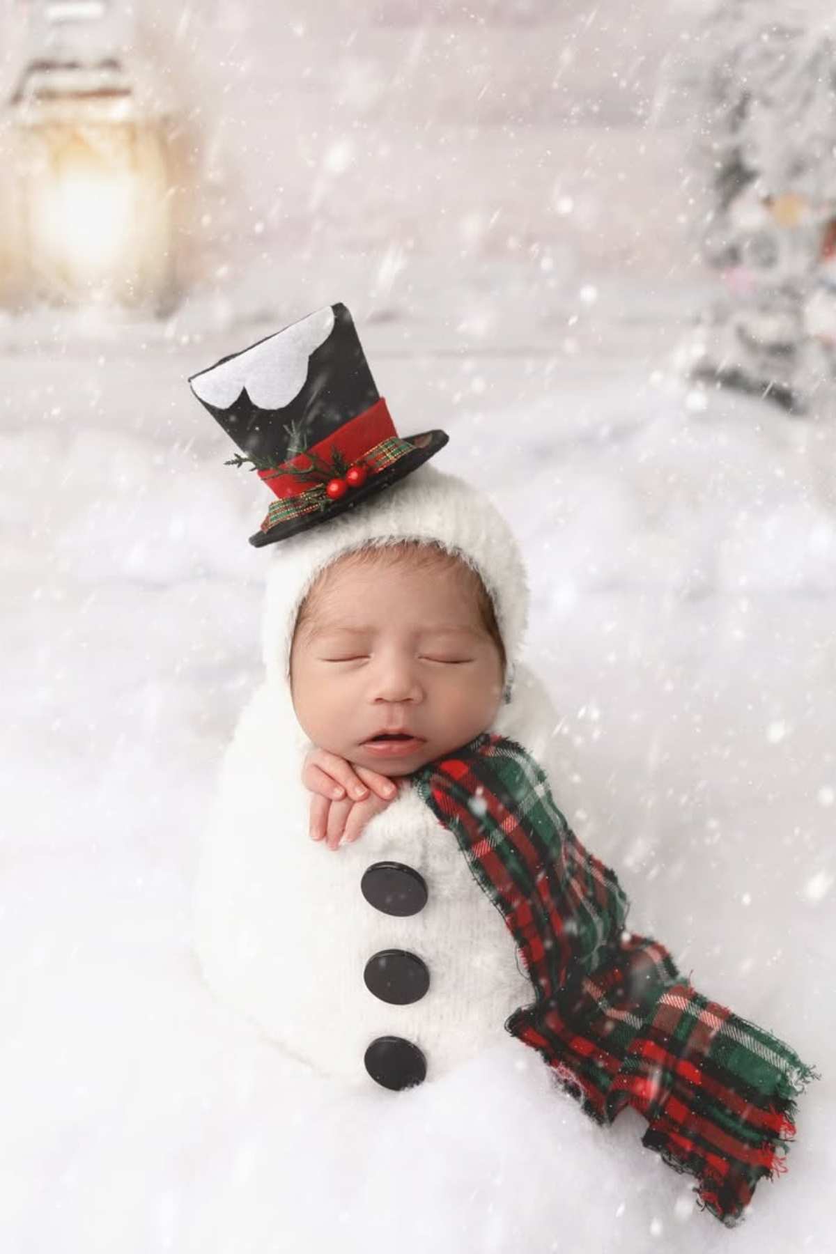A newborn baby is dressed as a snowman, wrapped in a white outfit with black buttons, a plaid scarf, and a small top hat, surrounded by artificial snow.