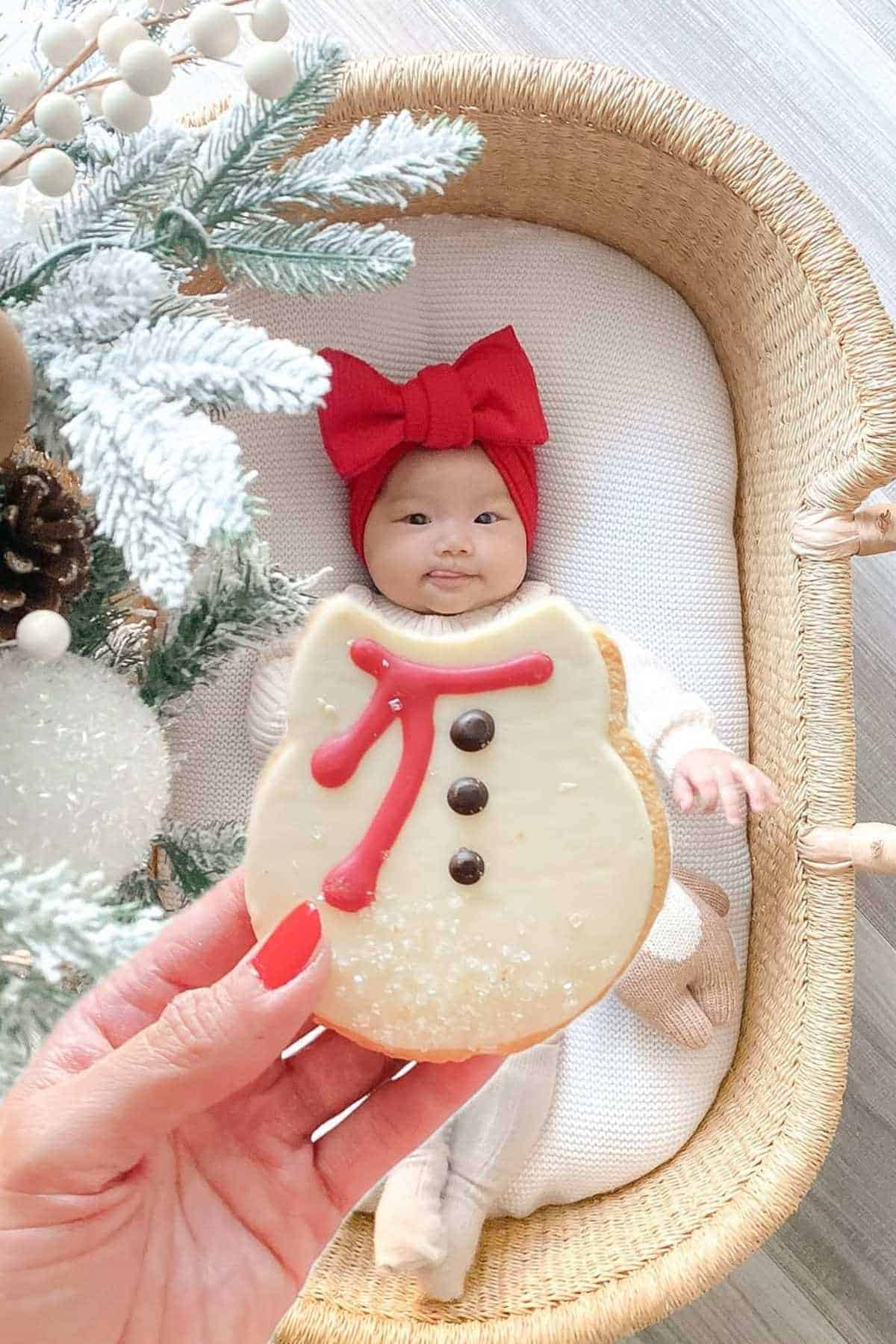 A baby wearing a red bow headband lies in a woven bassinet, while a hand holds a snowman-shaped cookie in the foreground.