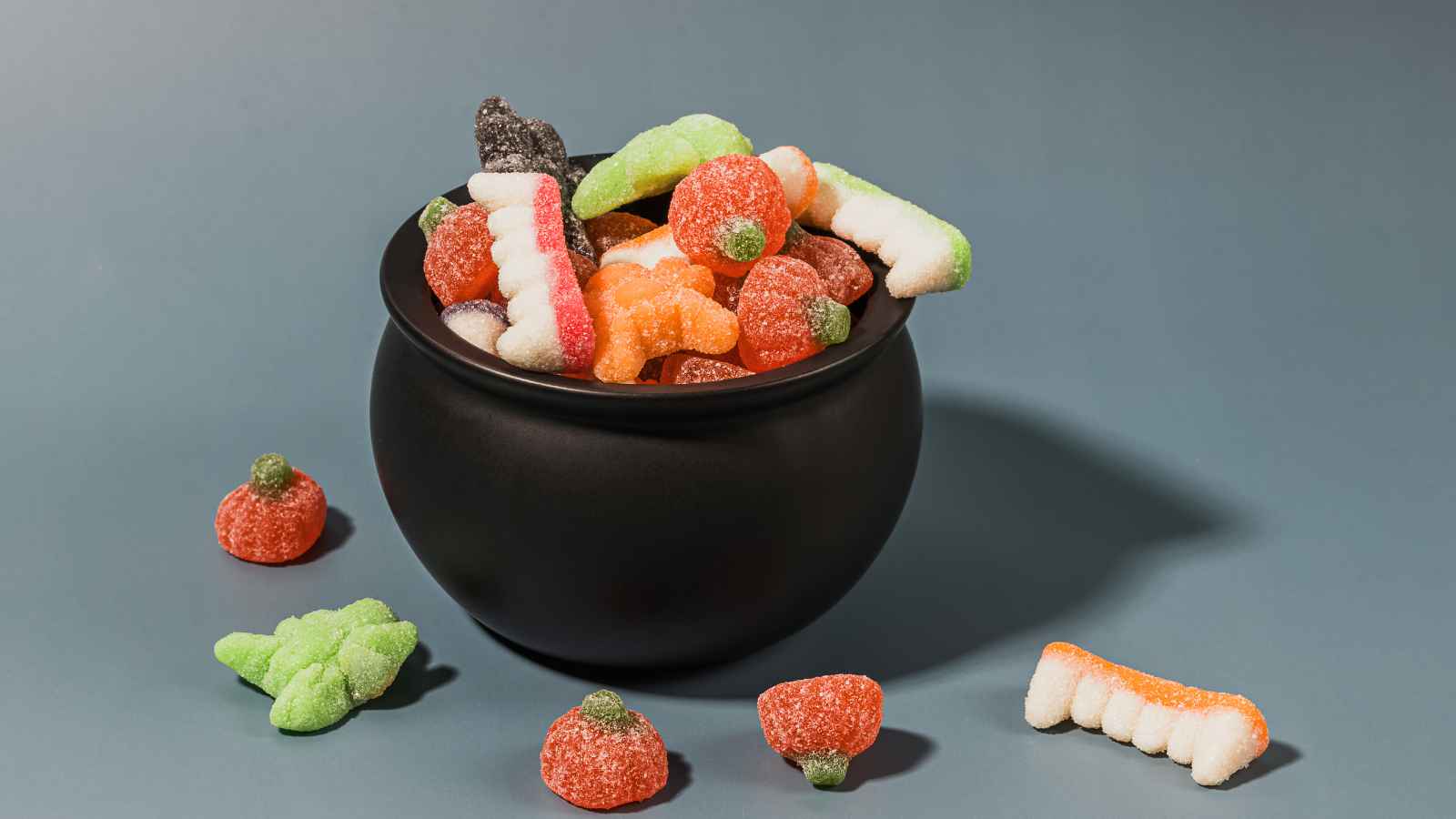 A black cauldron filled with colorful gummy candies shaped like pumpkins, bones, and other Halloween-themed figures, with some candies scattered around.