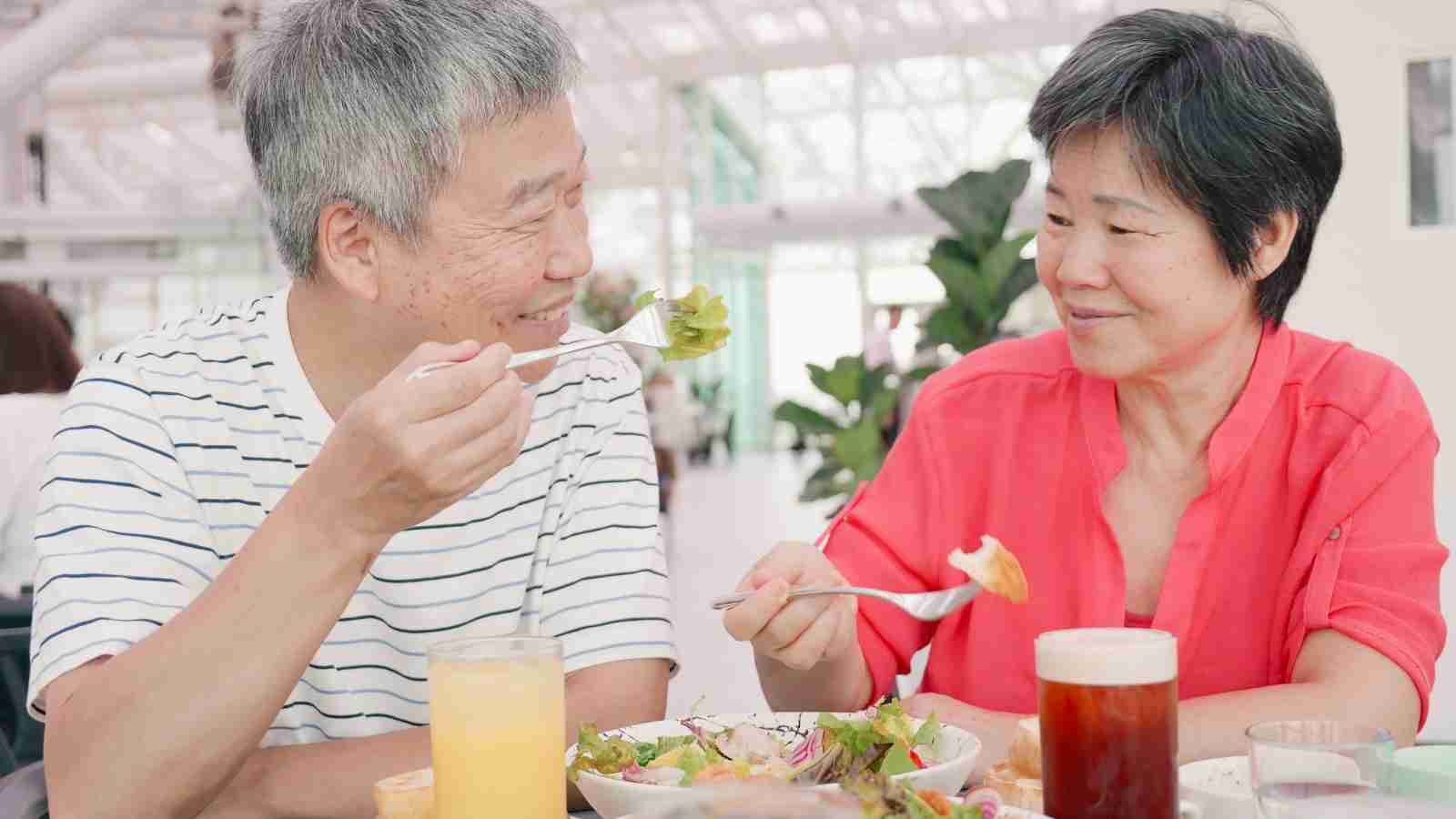 Two older adults sit at a table, smiling and eating salads with drinks in front of them in a bright indoor setting.
