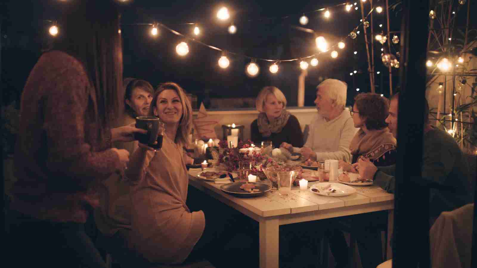 A group of people sit around a table outdoors at night, eating, drinking, and talking under string lights and candles.