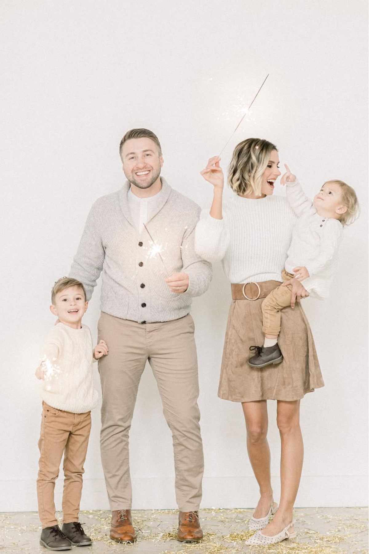 A family of four, dressed in light neutral clothing, stands together indoors, holding sparklers and smiling.