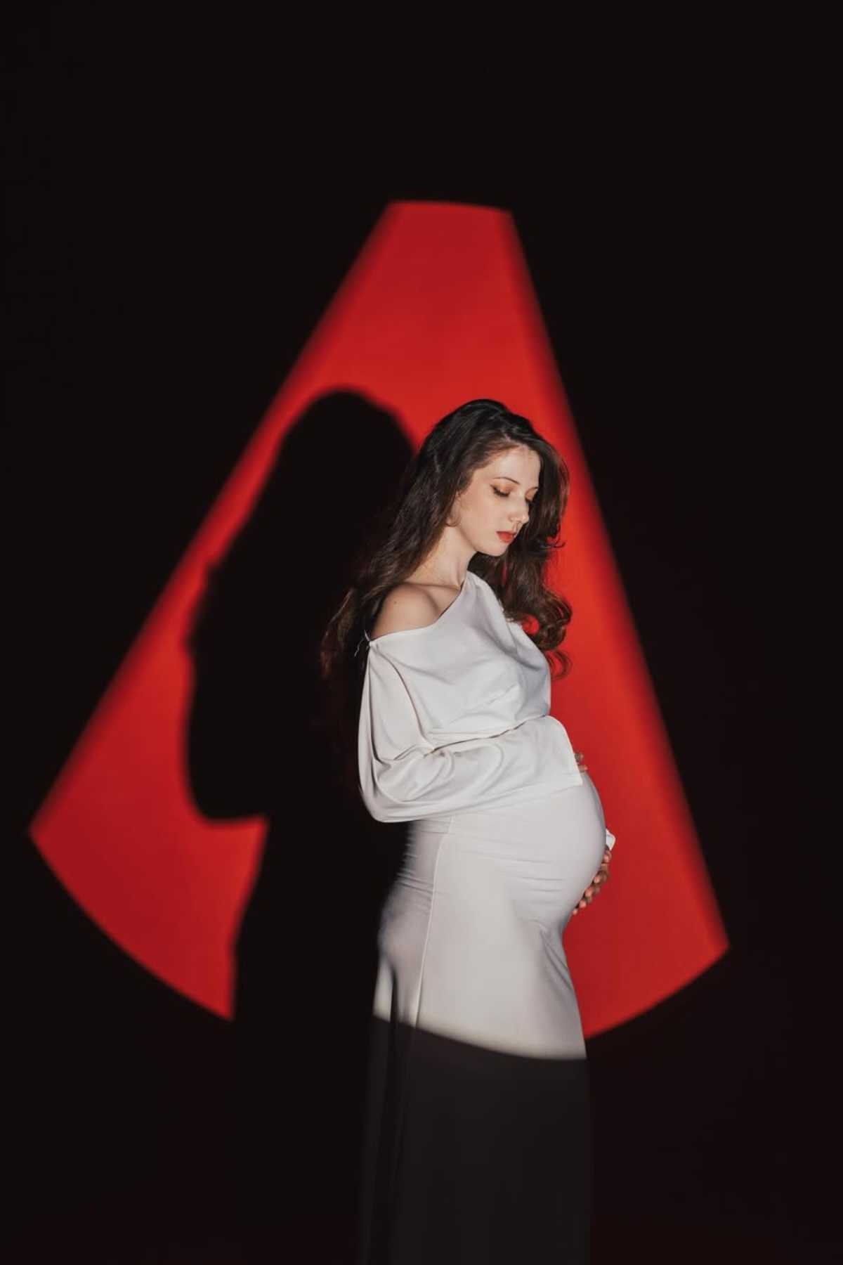 A pregnant woman in a white dress stands in profile, cradling her belly, with a dramatic red and black background and a shadow cast behind her.