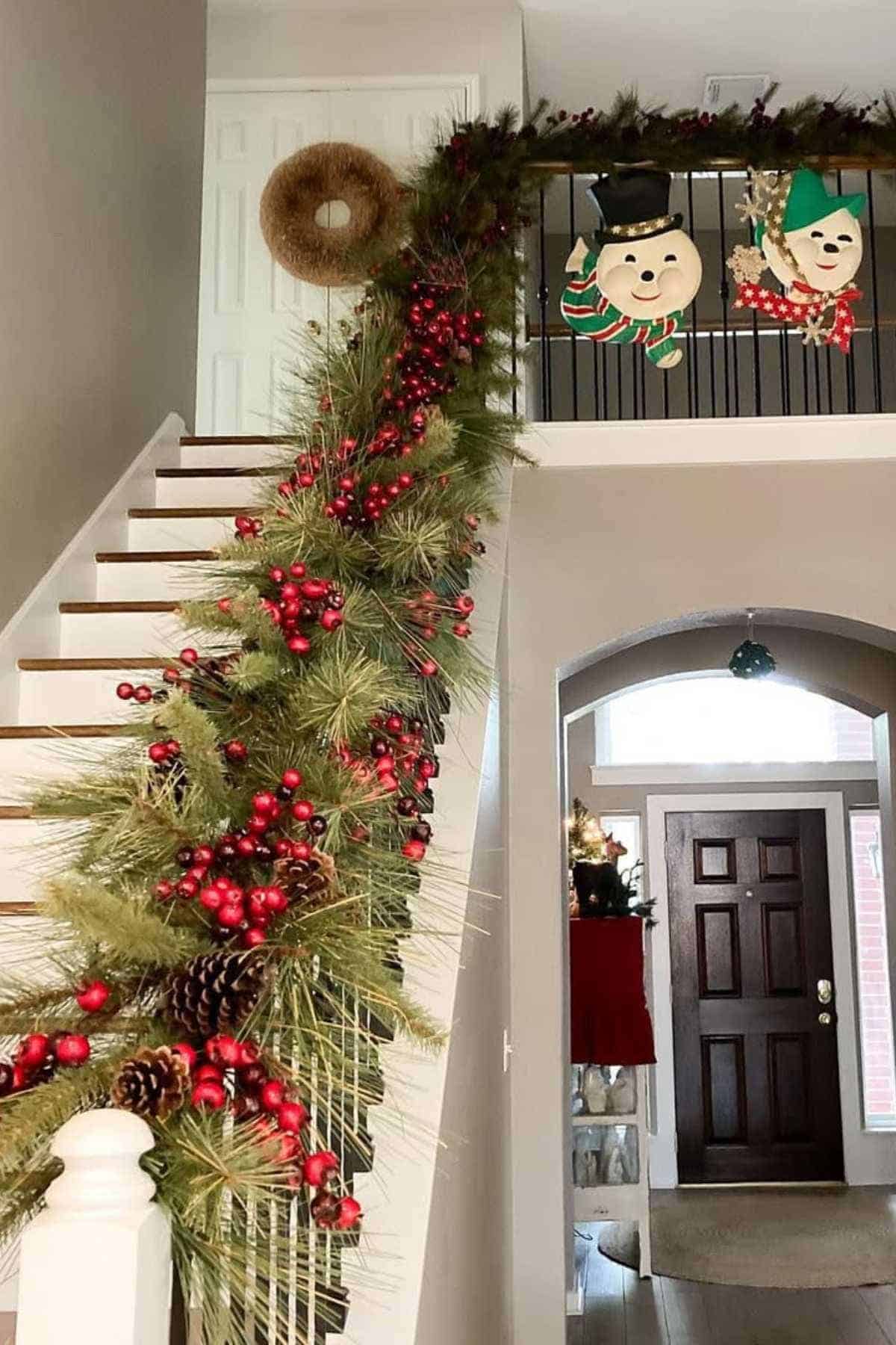A staircase decorated with garland, pinecones, and red berries, with wreaths and snowman faces hanging on the upper railing in a home entryway.