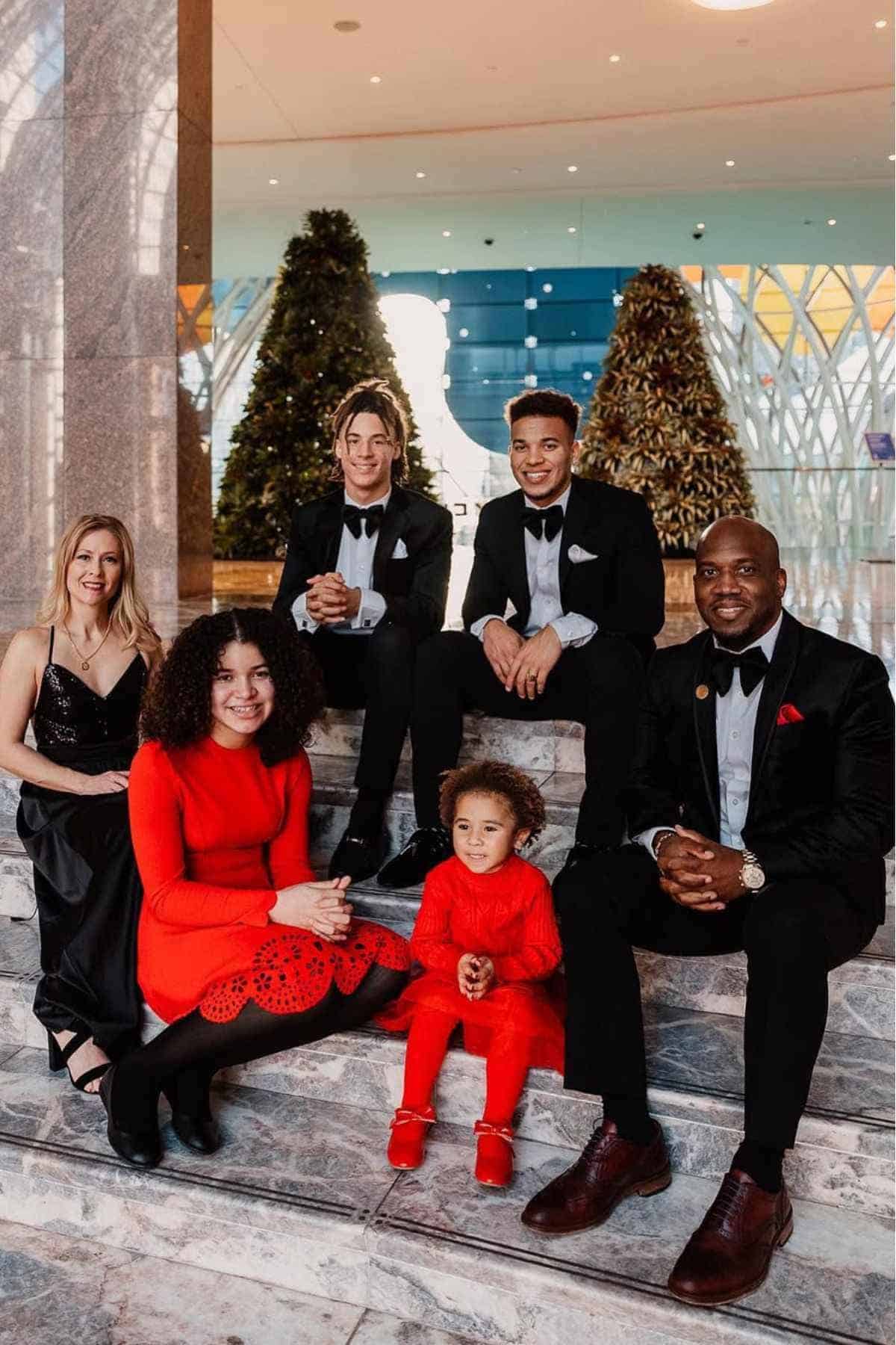 Six people dressed formally sit on marble steps in front of two decorated Christmas trees; three men in suits and ties, two women in dresses, and a young child in red.