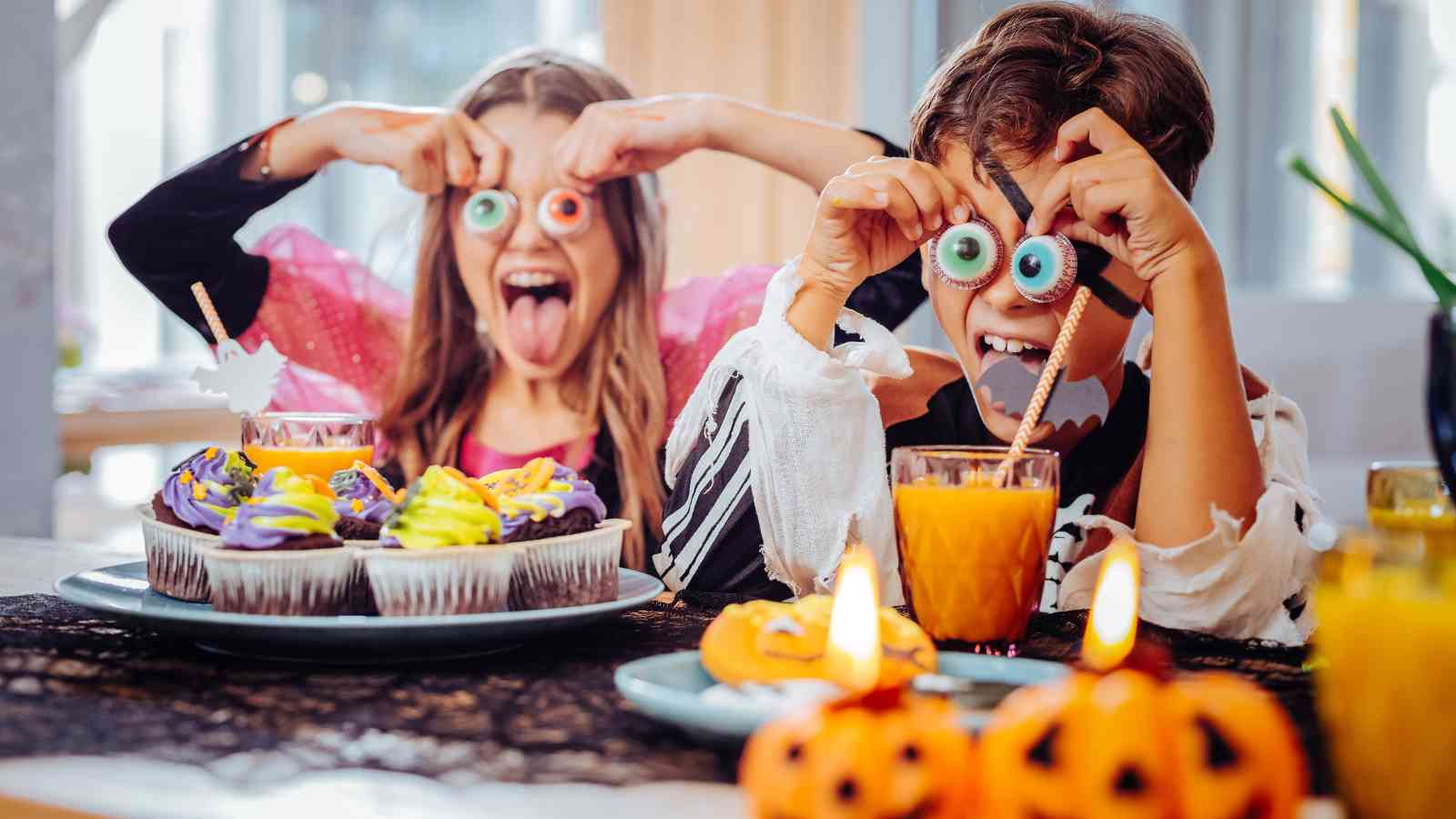 Two children in Halloween costumes hold fake eyeballs to their faces and make funny expressions at a decorated table with cupcakes, orange drinks, and small pumpkin decorations.