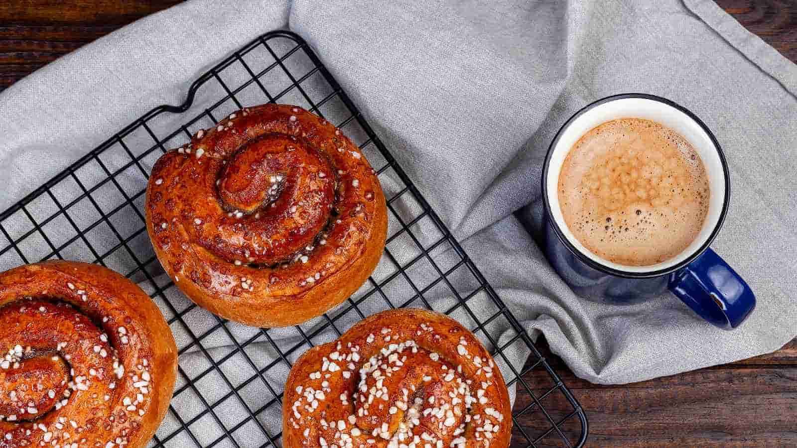 Three cinnamon buns with pearl sugar on a cooling rack next to a cup of coffee on a gray cloth.