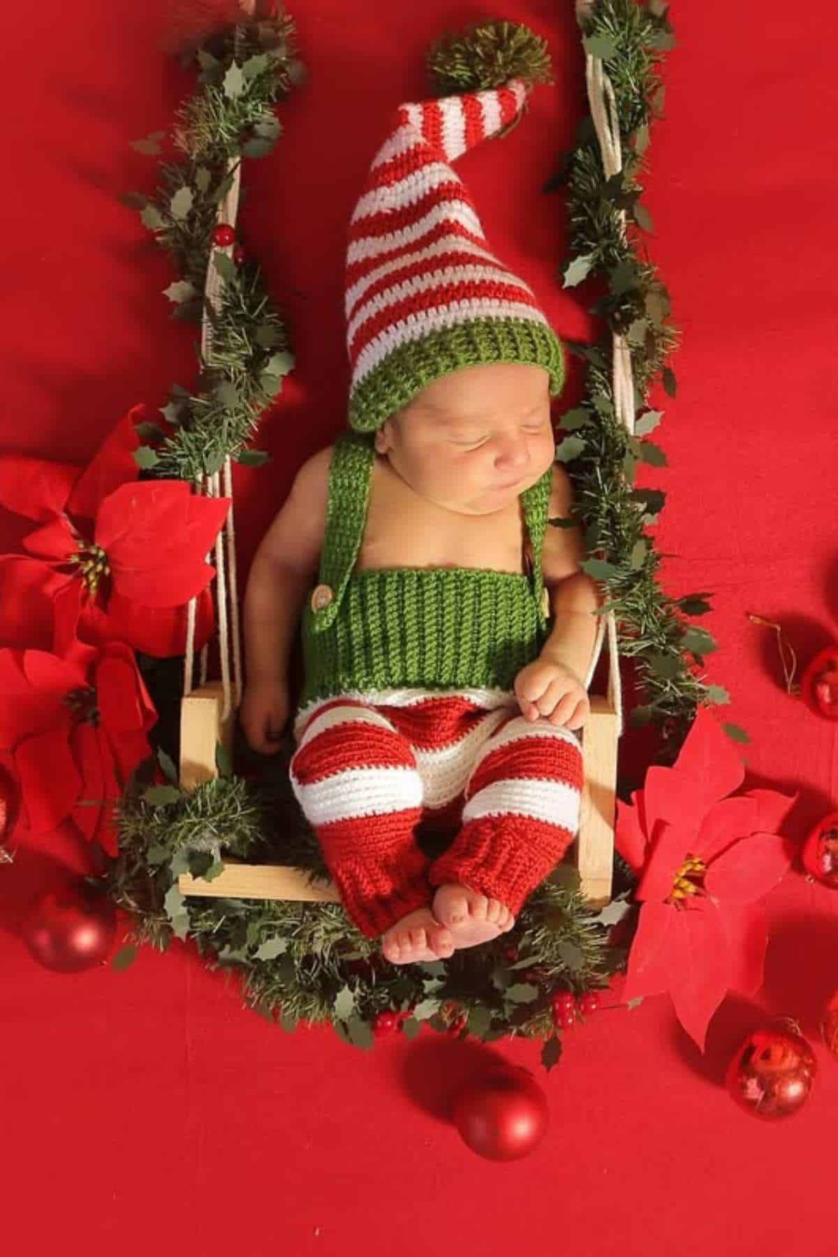 A newborn baby dressed in a red, white, and green striped elf outfit sleeps on a decorated swing surrounded by poinsettias and ornaments on a red background.