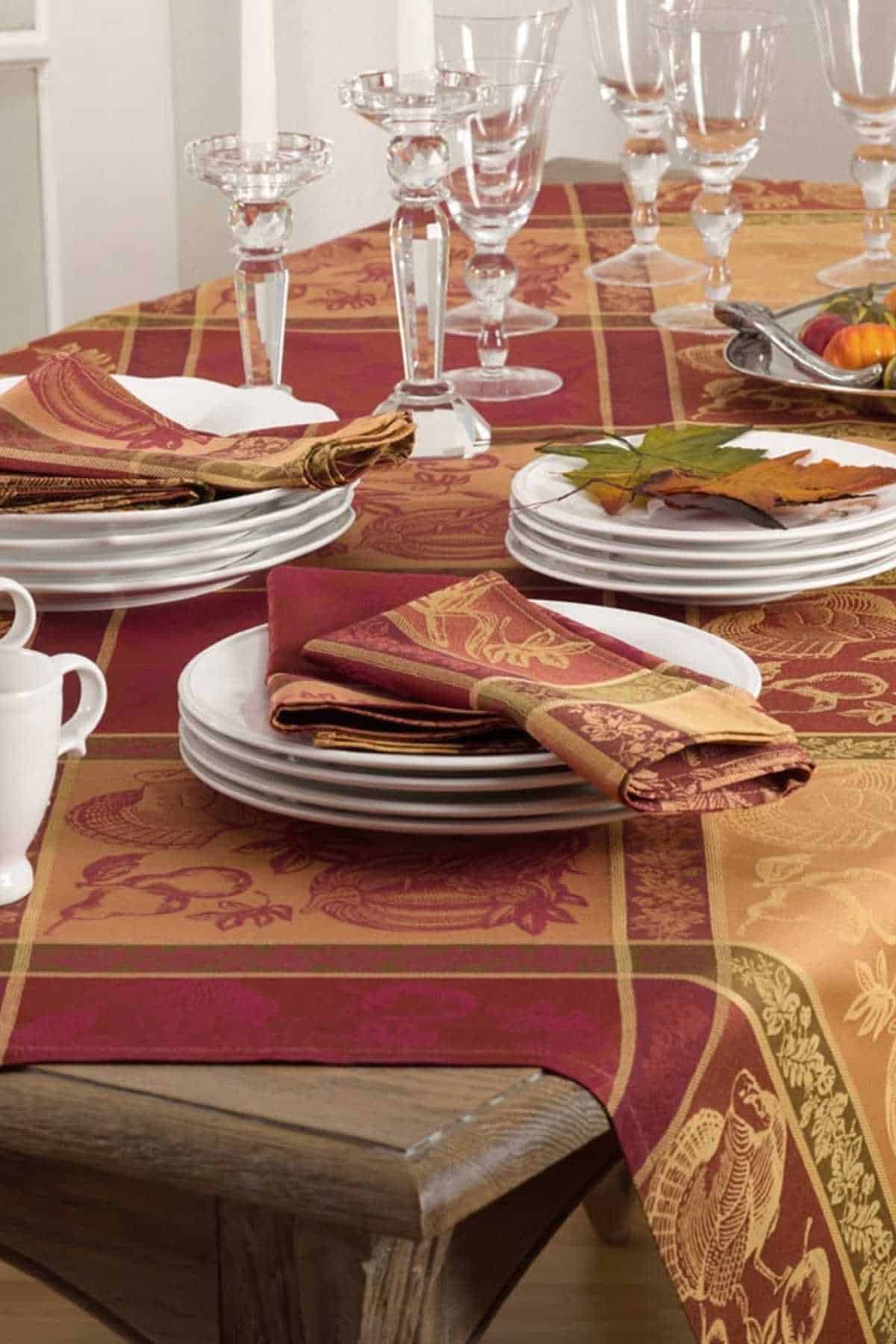 A dining table set with stacked white plates, folded red and gold napkins, clear glassware, and a red-orange patterned tablecloth with a fall theme.