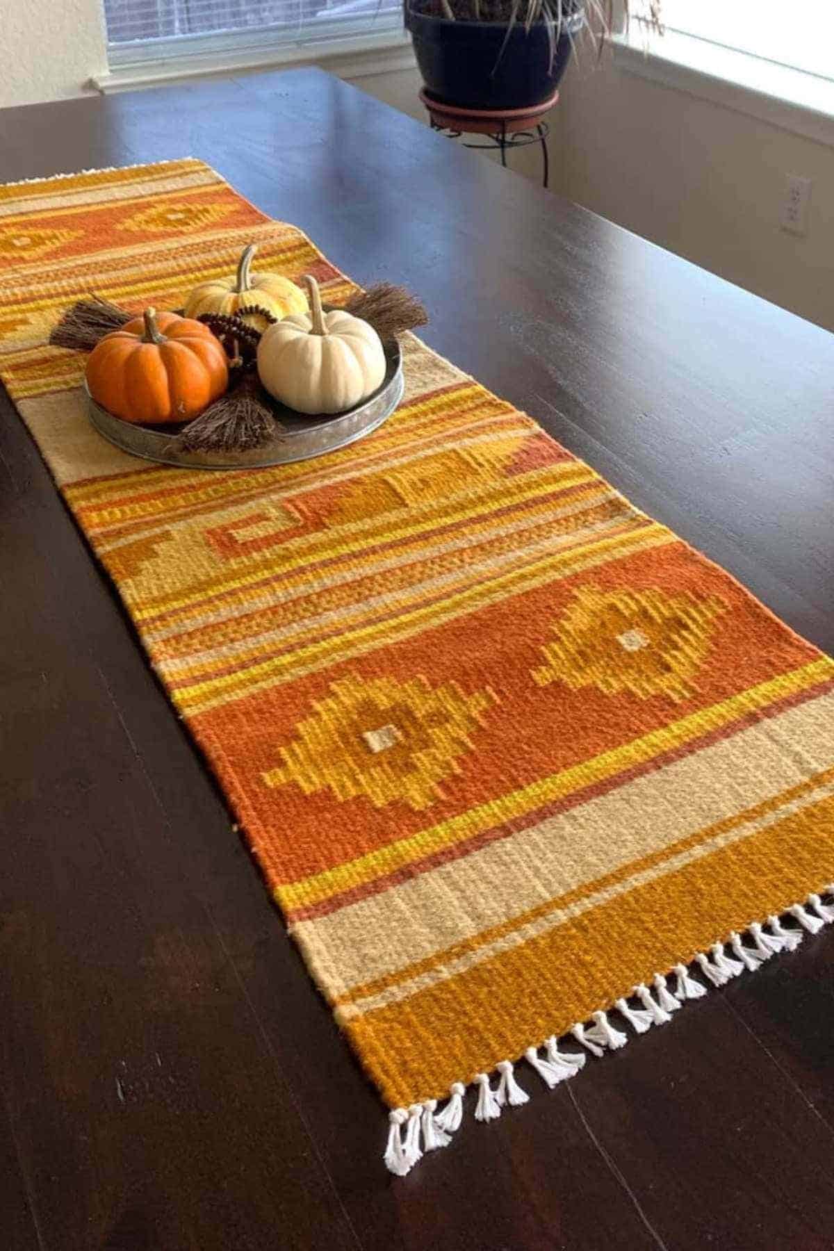 A dark wooden table with a yellow and orange patterned woven table runner, topped with a metal tray holding pumpkins and decorative brooms.