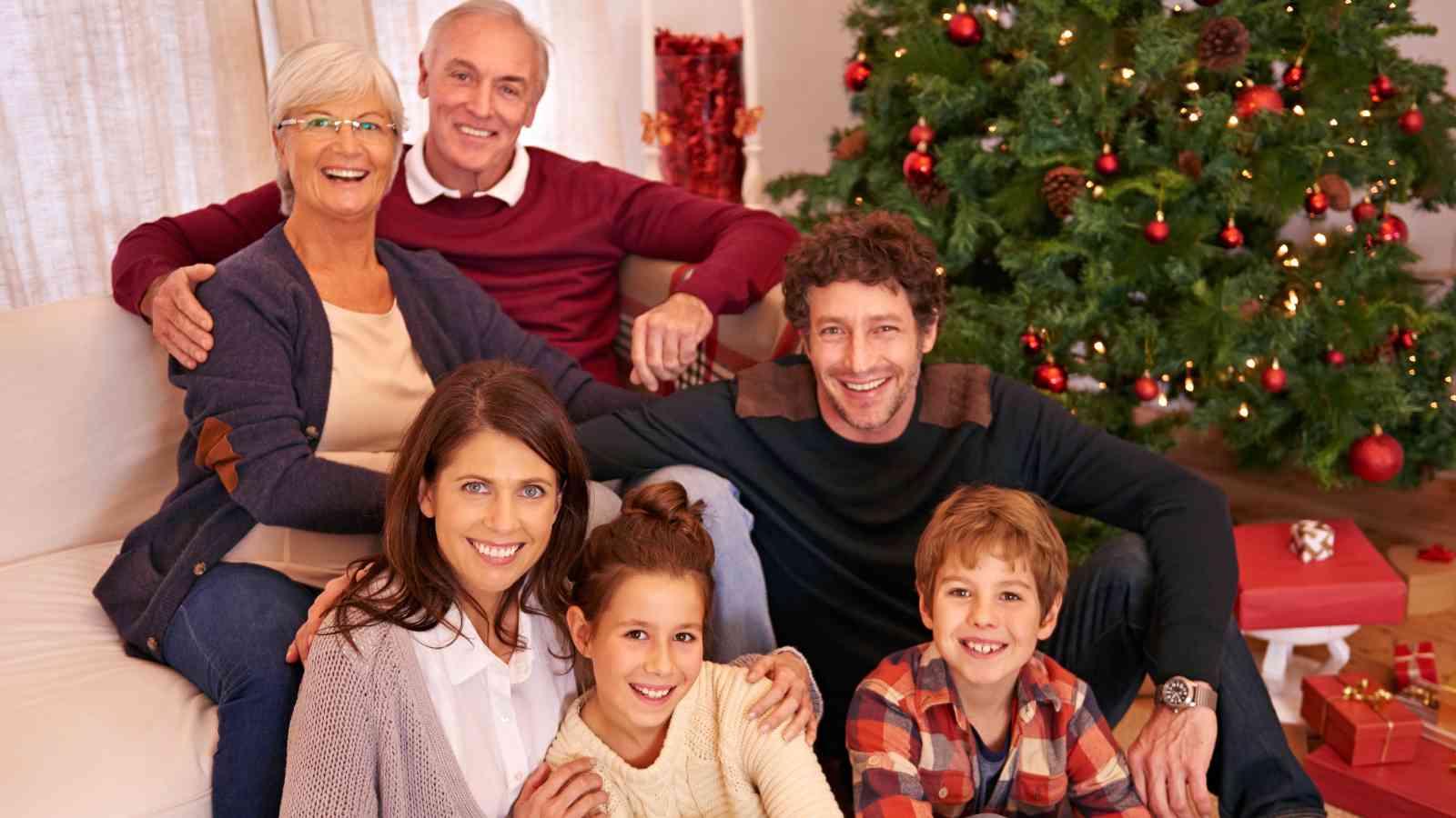 Three adults and two children sit and smile in front of a decorated Christmas tree, with gifts visible under the tree, in a warmly lit living room.