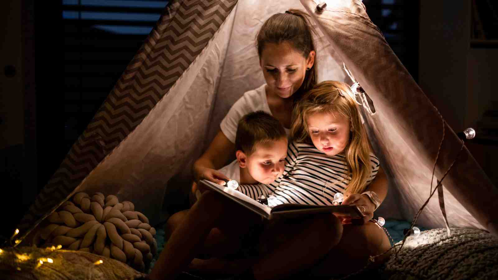 A woman sits inside a tent indoors, reading a book to two young children under warm string lights.