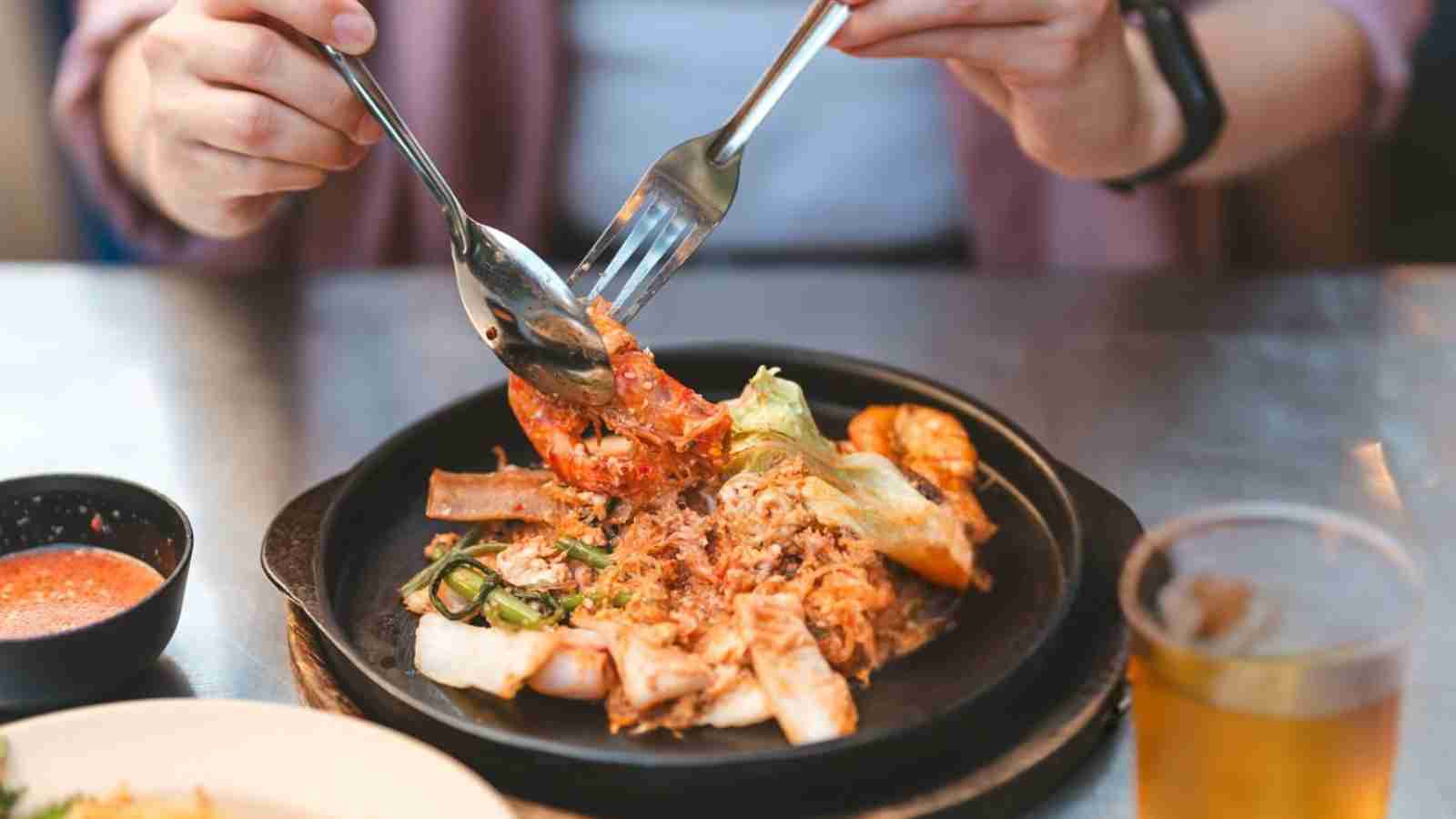 A person uses a fork and spoon to eat a dish with shrimp and vegetables on a black plate, with a glass of drink and a bowl of sauce nearby.