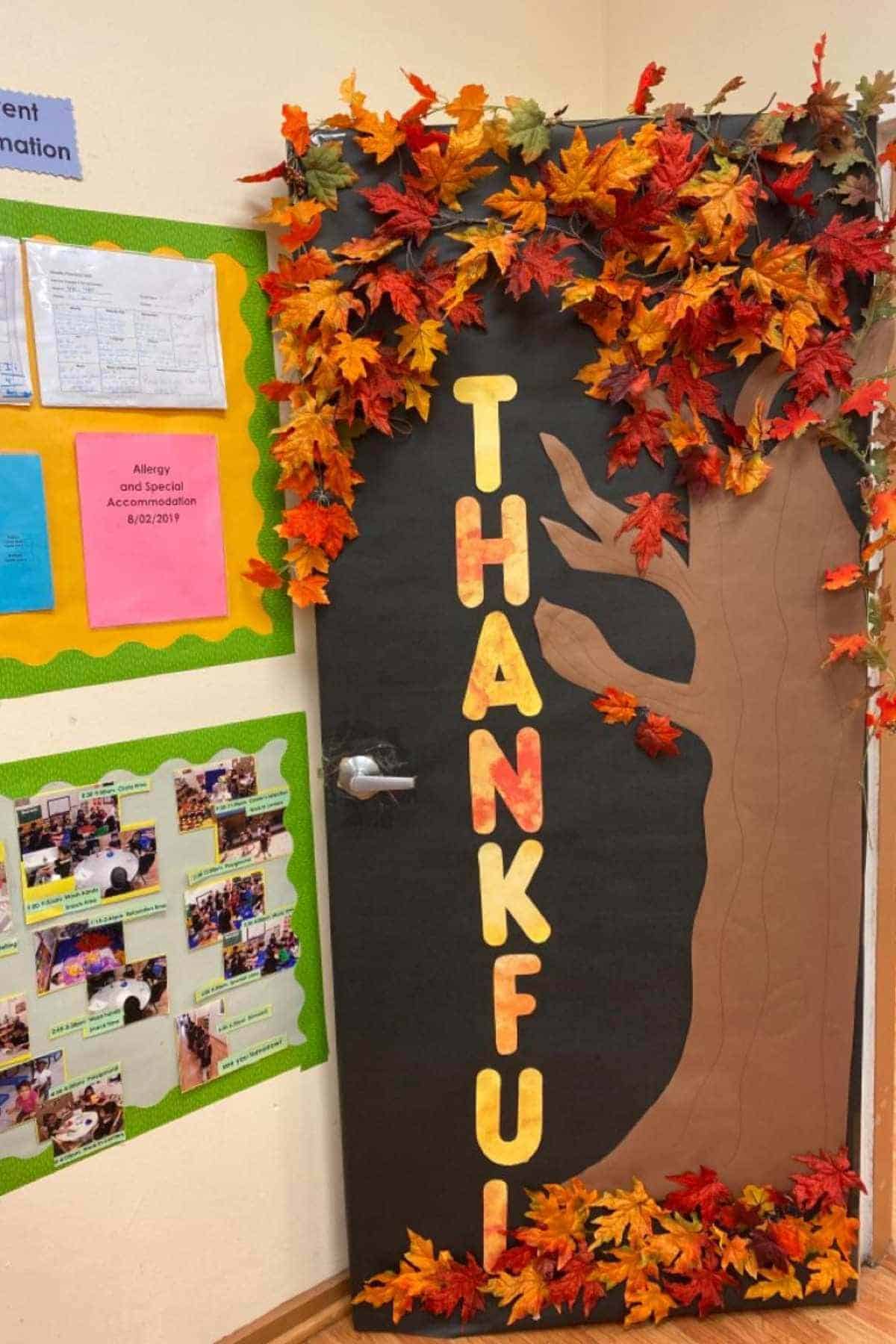 A classroom door decorated with a tree, autumn leaves, and the word "THANKFUL" spelled vertically in large letters. Displays with papers and photos are on the adjacent wall.