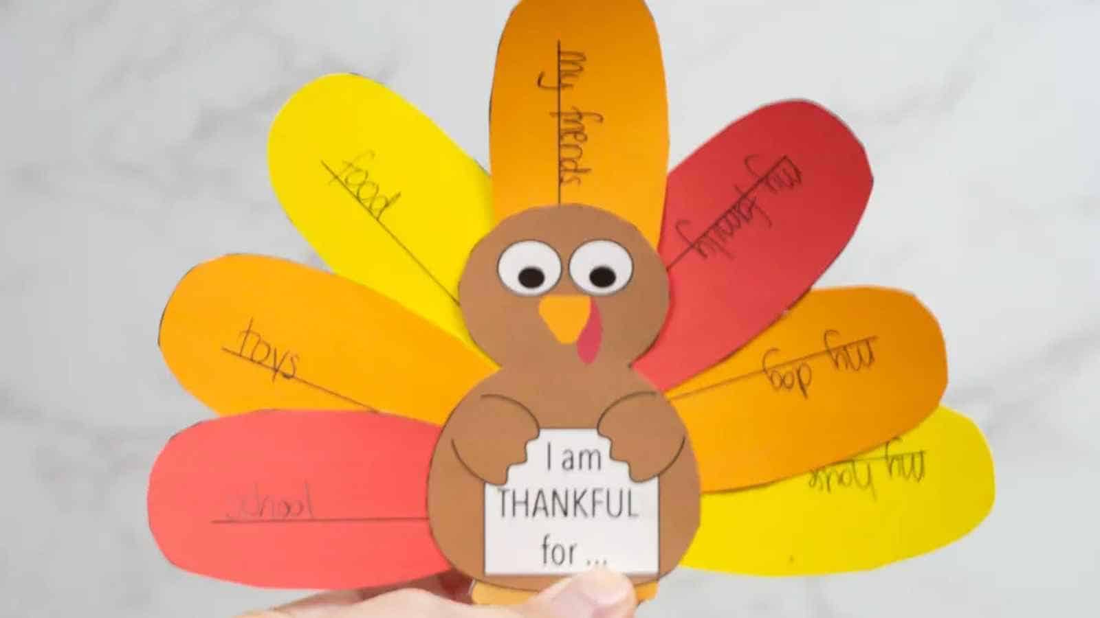 A hand holds a paper turkey craft with colored feathers labeled with things to be thankful for, such as food, toys, and friends. The turkey holds a sign that says