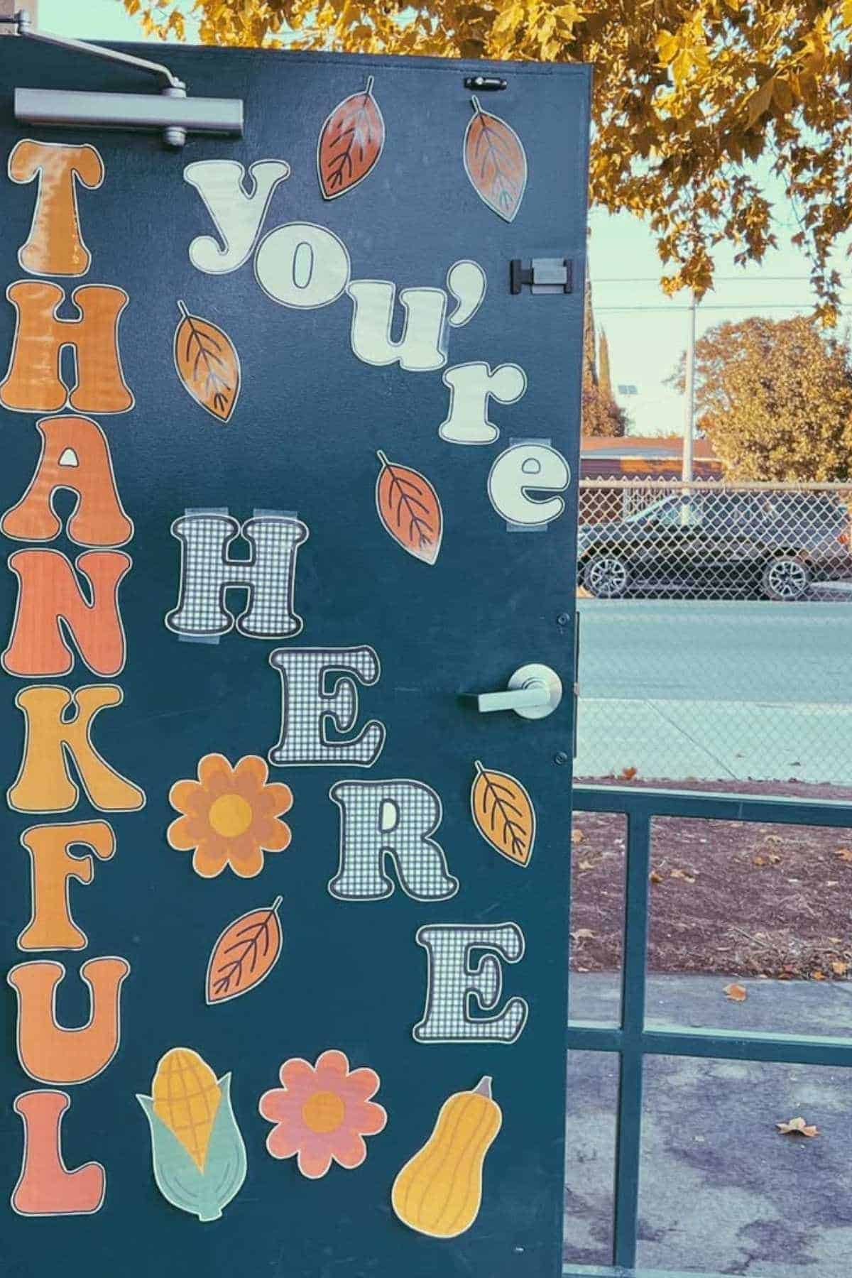 A door decorated with the words "THANKFUL you’re HERE," colorful letters, leaves, flowers, corn, and a gourd, with a parking lot and trees visible in the background.